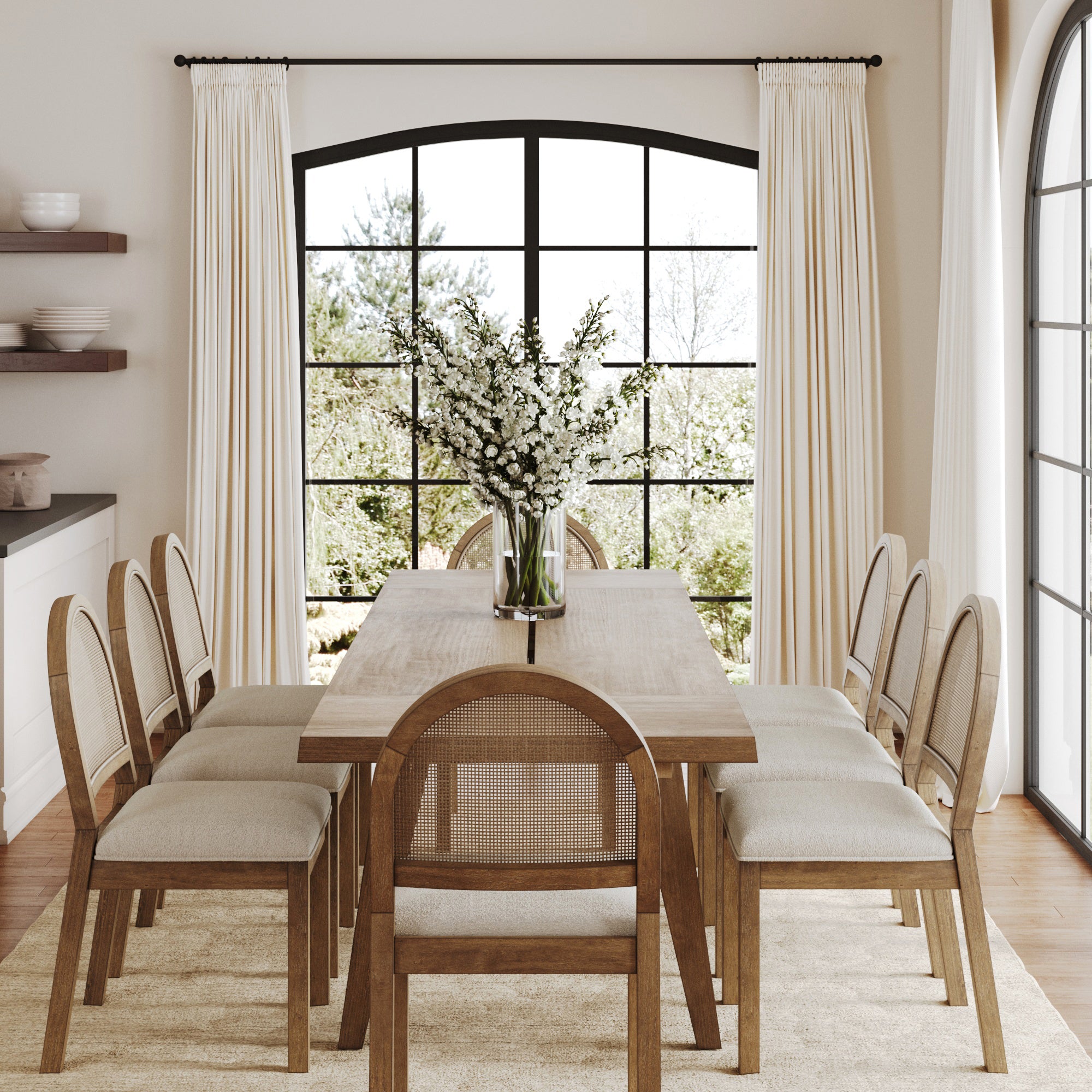 Bright dining room featuring the Nathan James Natural Rattan Dining Table Set (8 Chairs), a vase of white flowers centerpiece, large black-framed windows with white curtains, neutral rug on wood floors, and lush greenery outside.