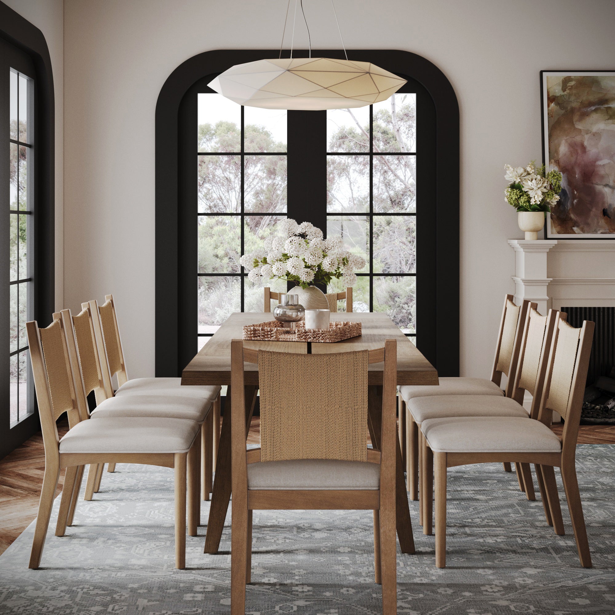 A modern dining room with the Nathan James Modern Farmhouse Table Extension in oak veneer light brown, solid legs, eight upholstered chairs, a white flower centerpiece, large windows with white curtains, and views of trees outside.