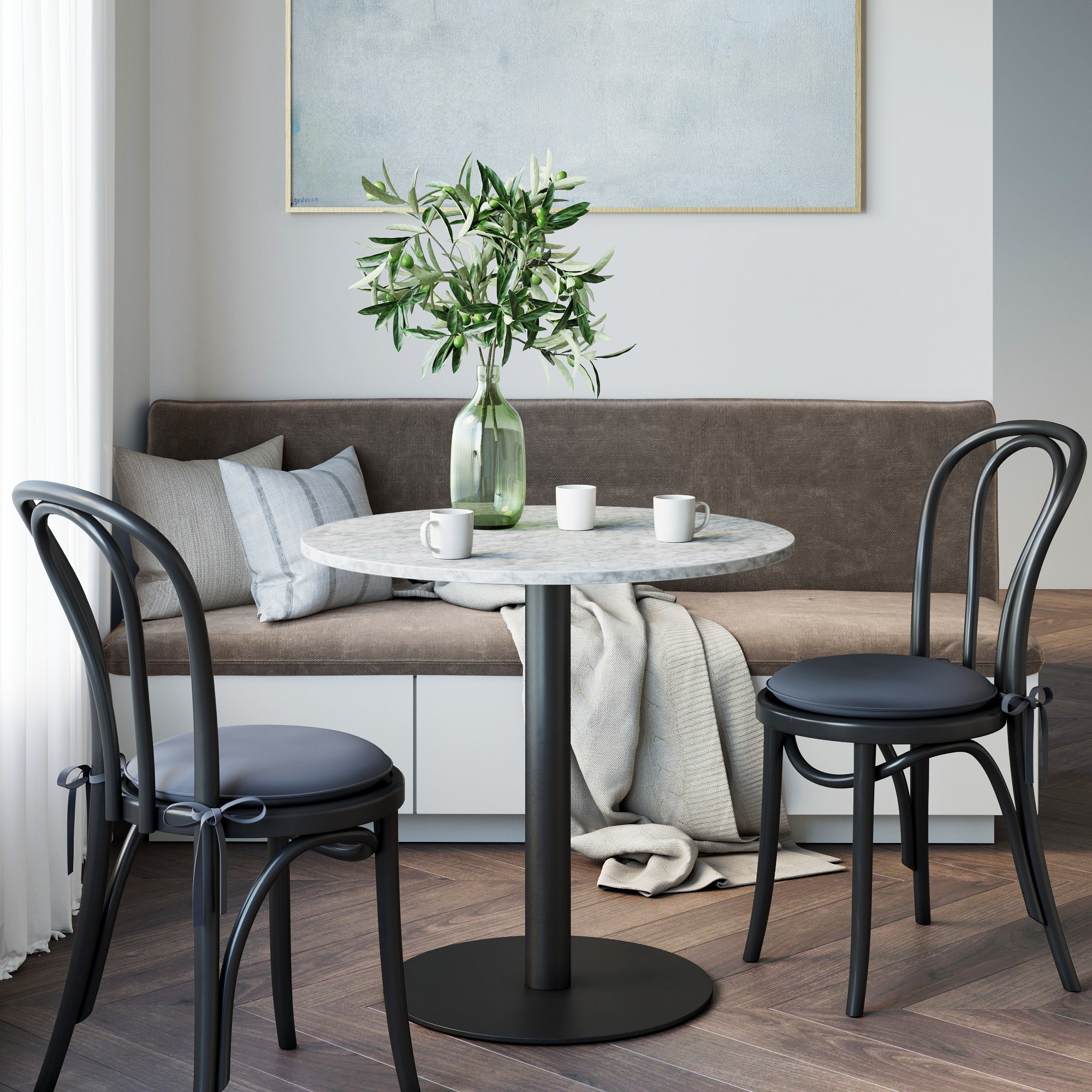 A Nathan James Faux Marble & Metal Bistro Dining Table in black with two chairs stands before a gray sofa topped with a glass vase of greenery and three white mugs; a beige throw drapes over the sofa, creating a cozy, modern vibe.