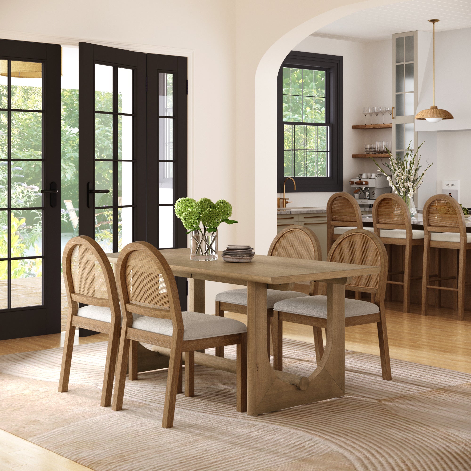 A modern dining room features the Nathan James Dining Table Set for 4 with light brown rattan chairs in boucle, a vase of green flowers, and large black-framed glass doors overlooking a garden. Light wood floors and neutral decor complete the look.