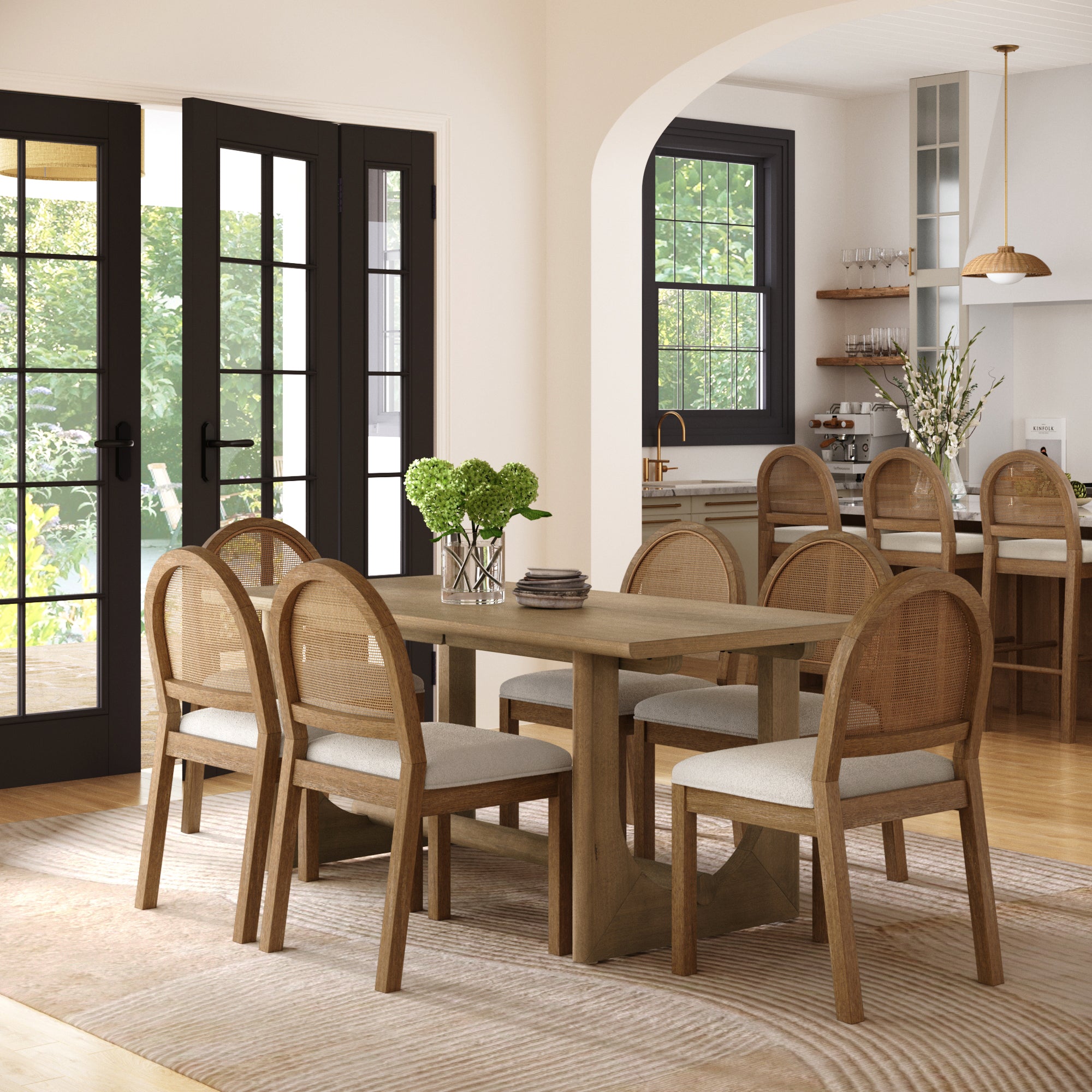A bright dining room showcases the Nathan James Dining Table Set for 6 with Rattan Dining Chairs in bouclé/light brown, accented by a vase of green flowers. Sunlight streams through large glass doors, revealing the kitchen beyond.