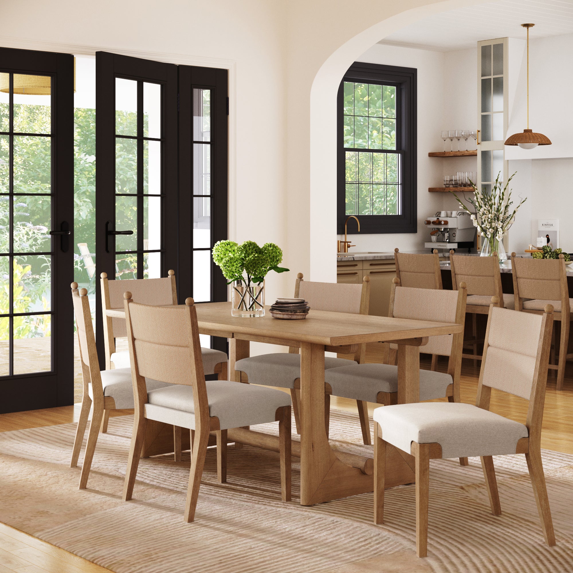 A modern dining room showcases the Nathan James Boho Dining Table Set for 6, featuring seagrass chairs, on a light rug. Large glass doors and an archway open to a bright kitchen with open shelves and a vase of flowers.