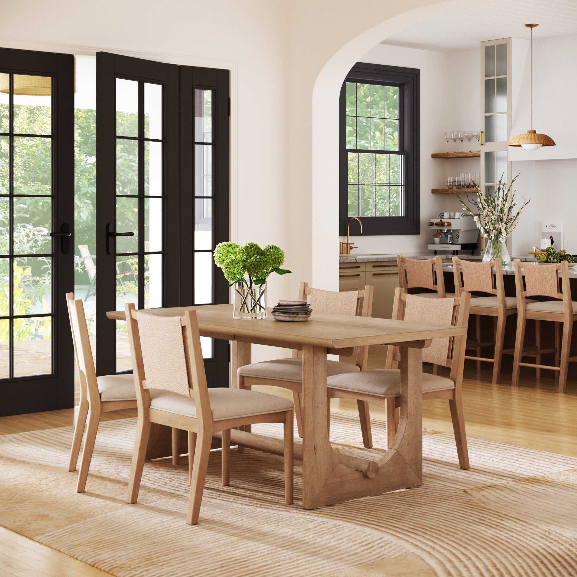 A bright dining room showcases the Nathan James Bohemian Dining Table Set with Upholstered Chairs for 4, centered on a striped rug. Large windows fill the space with natural light, and a kitchen with bar stools and pendant lights is visible behind.