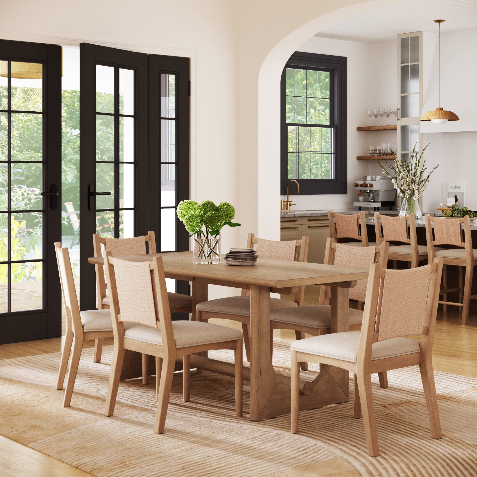 A bright dining room showcases the Nathan James Bohemian Dining Table Set with upholstered chairs for 6, a glass vase with green hydrangeas centerpiece, large windows, and an open view into a kitchen with shelves and gold pendant lights.