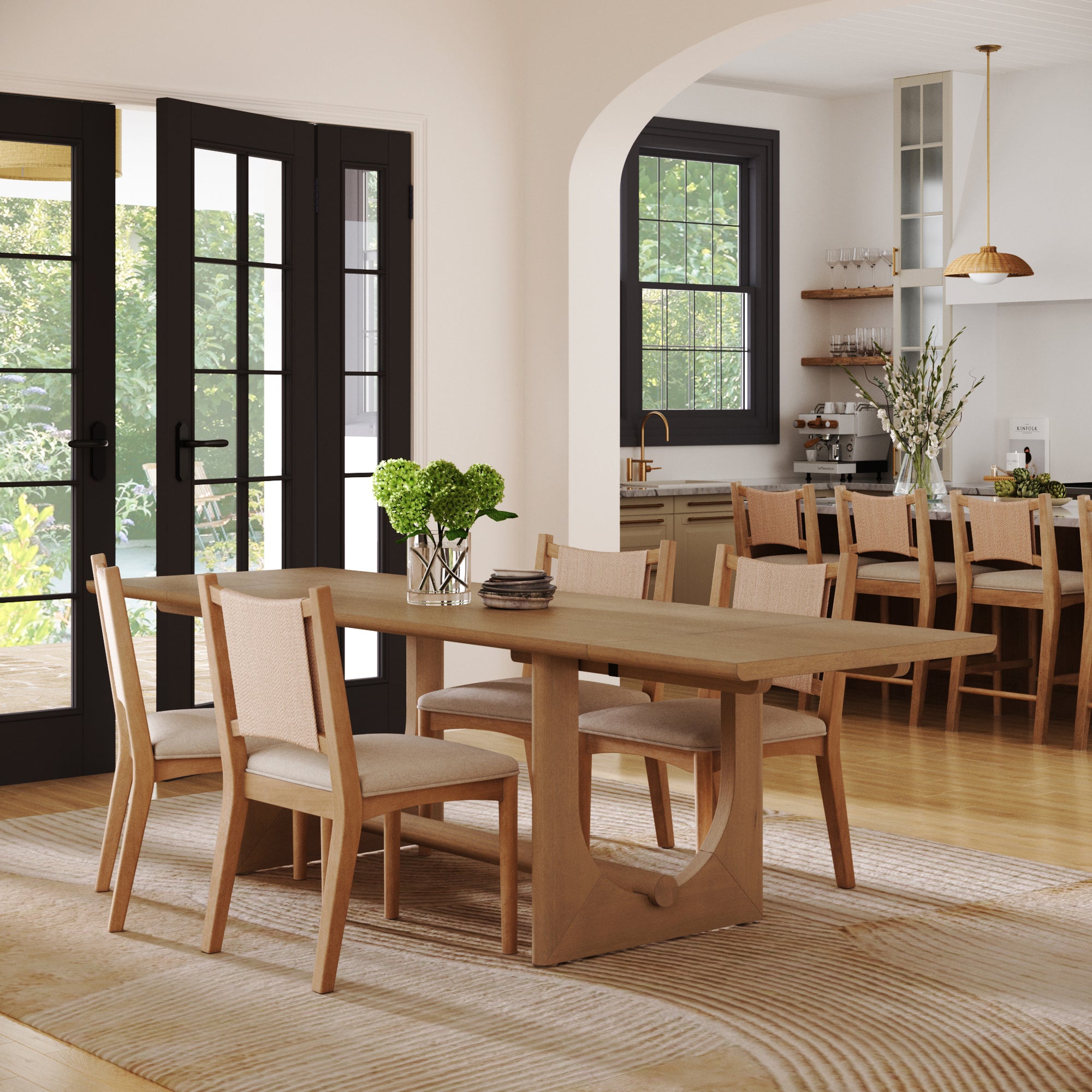A bright, modern dining room showcases the Nathan James Extendable Dining Table Set with 4 upholstered chairs, a vase of green hydrangeas, large windows, and an open view to a kitchen with wooden shelves and pendant lighting.