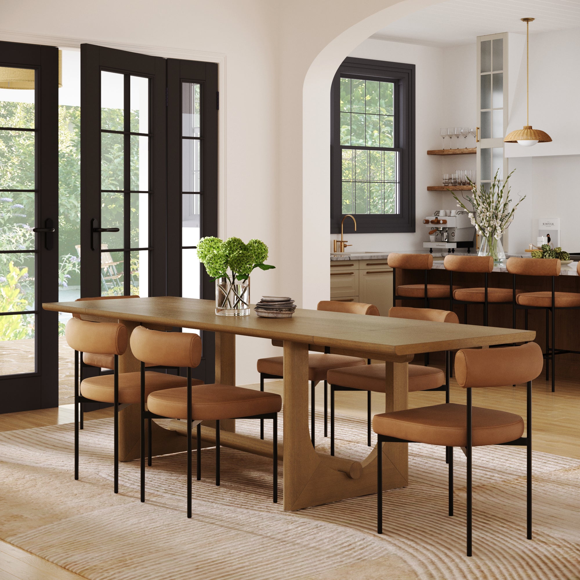 A modern dining room features the Nathan James Extendable Dining Table Set with 6 faux leather chairs, a vase of green flowers, large windows, and an open view to a kitchen with shelves, pendant lights, and neutral decor.