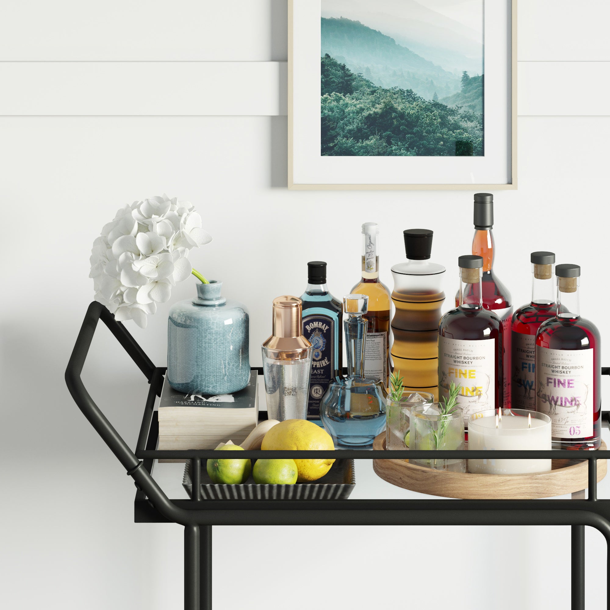 The Nathan James Metal Rolling Bar Cart features tempered glass shelves and a sleek black frame, displaying liquor bottles, mixers, flowers, lemons, limes, candles, and a blue ceramic jar below a framed landscape on a white wall.