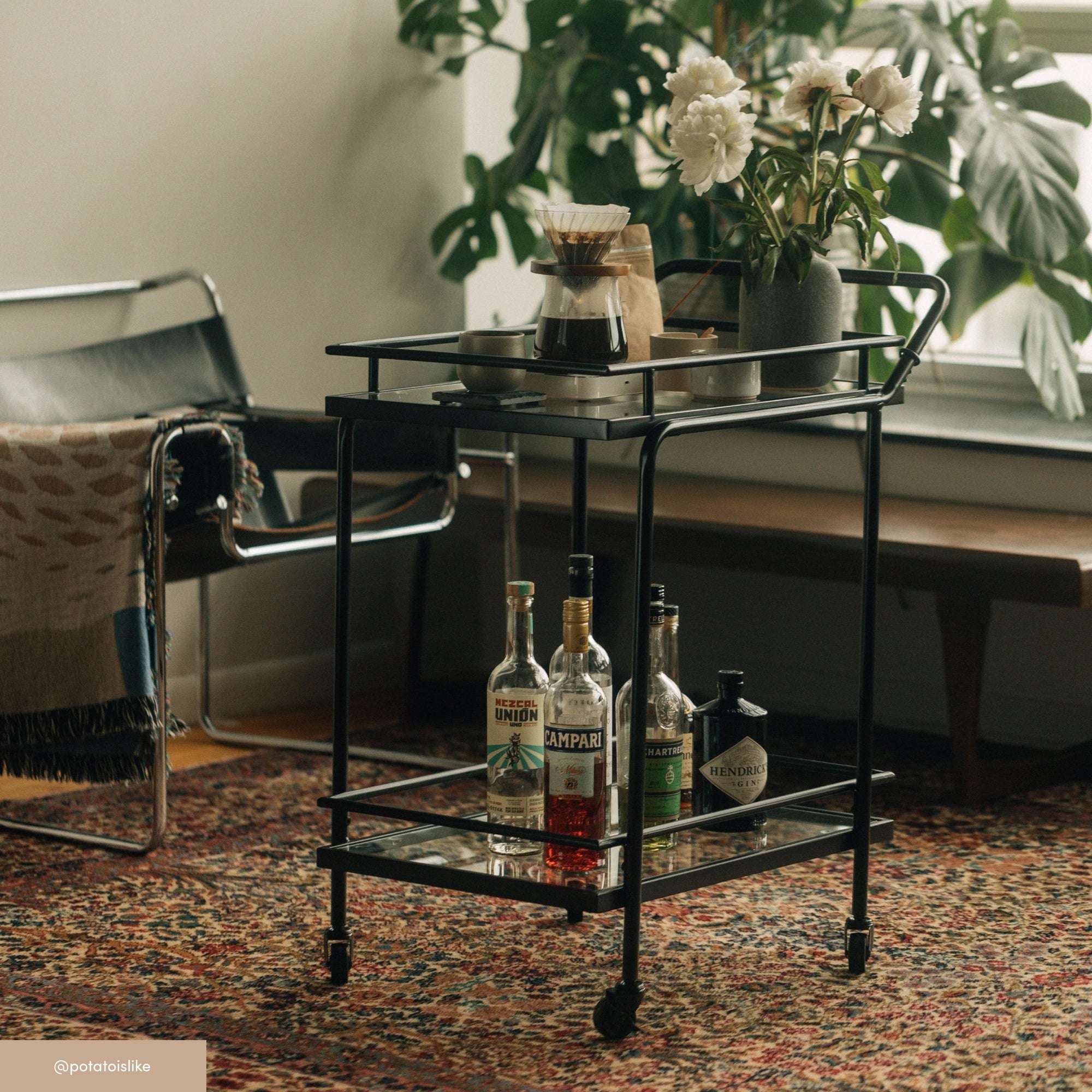 A black bar cart with bottles of liquor on the lower shelf and coffee-making items and a vase of white flowers on top, sits on a patterned rug next to a chair and a large leafy plant by a window.