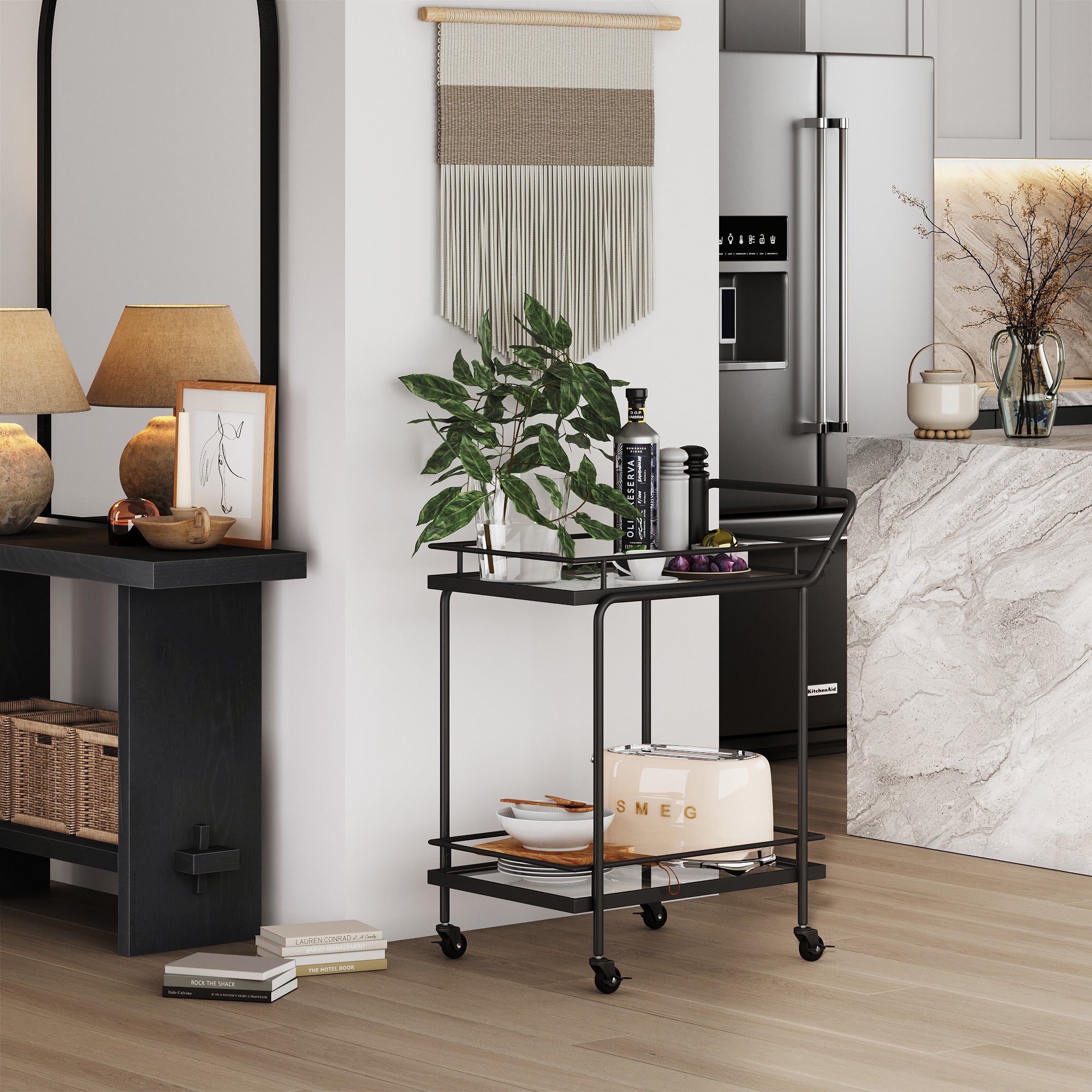 A modern kitchen features a black metal bar cart with plants, bottles, and a white SMEG toaster. The background shows a marble countertop, stainless steel fridge, and neutral decor with lamps and woven baskets.