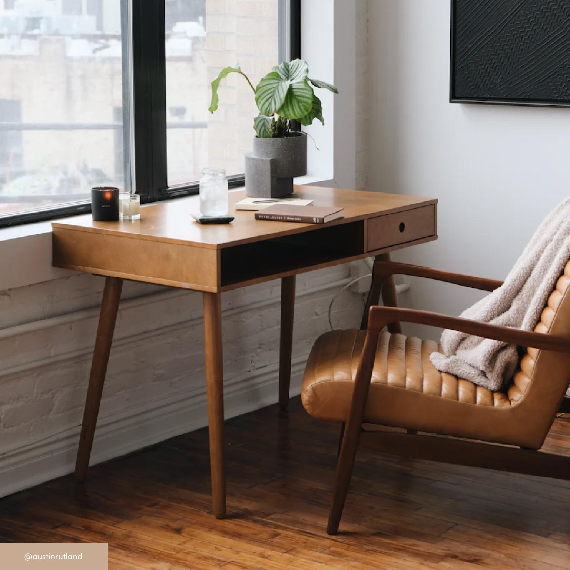 A mid-century modern desk with a potted plant, notebook, candle, and glass sits by a large window. A tan leather chair with a blanket is placed next to the desk on a wooden floor.