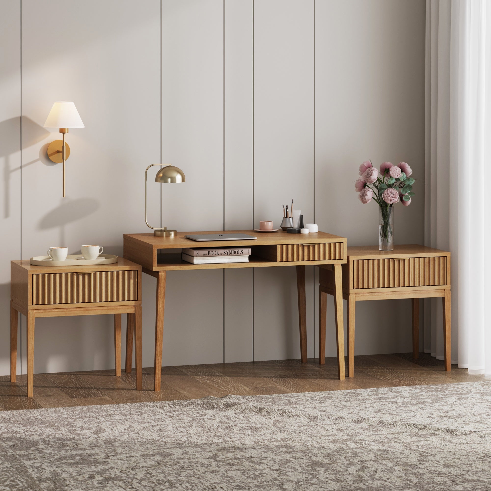 The Nathan James Solid Wood Fluted Desk & 2 Nightstands Bedroom Vanity Set stands against a light-paneled wall, adorned with a gold lamp, books, small decor, a wall-mounted lamp, and a vase of pink flowers for a Mid-Century Modern touch.