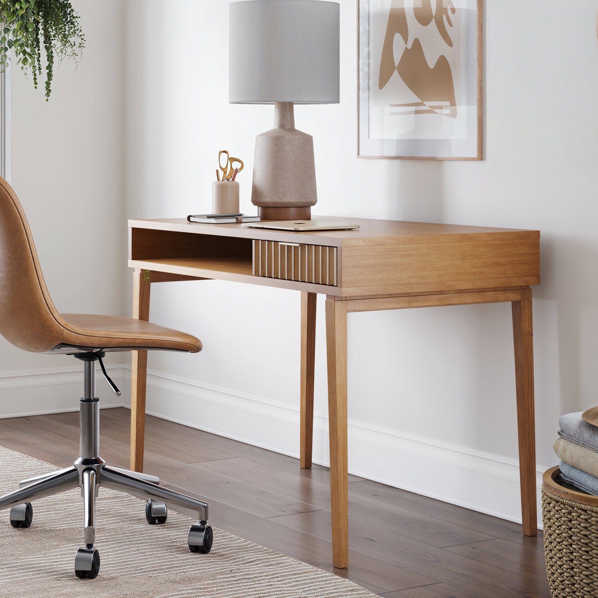 The Nathan James Mid-Century Modern Fluted Desk with Drawer features a tan wheeled chair, beige lamp, and gold accents. Set against a white wall with minimalist art and a hanging plant, it creates a chic bohemian workspace.