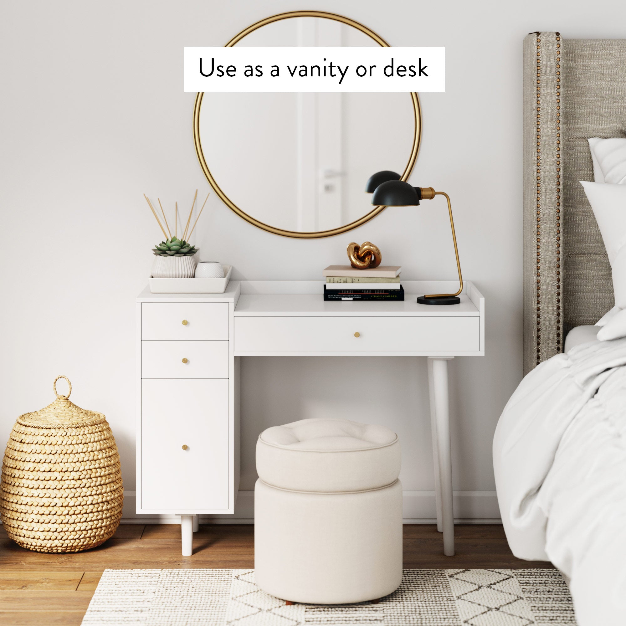 A modern white vanity or desk with drawers stands against a wall, topped with a lamp and decor. A round gold-framed mirror hangs above, with a cushioned stool below. A woven basket and bed are nearby in a cozy, bright bedroom.
