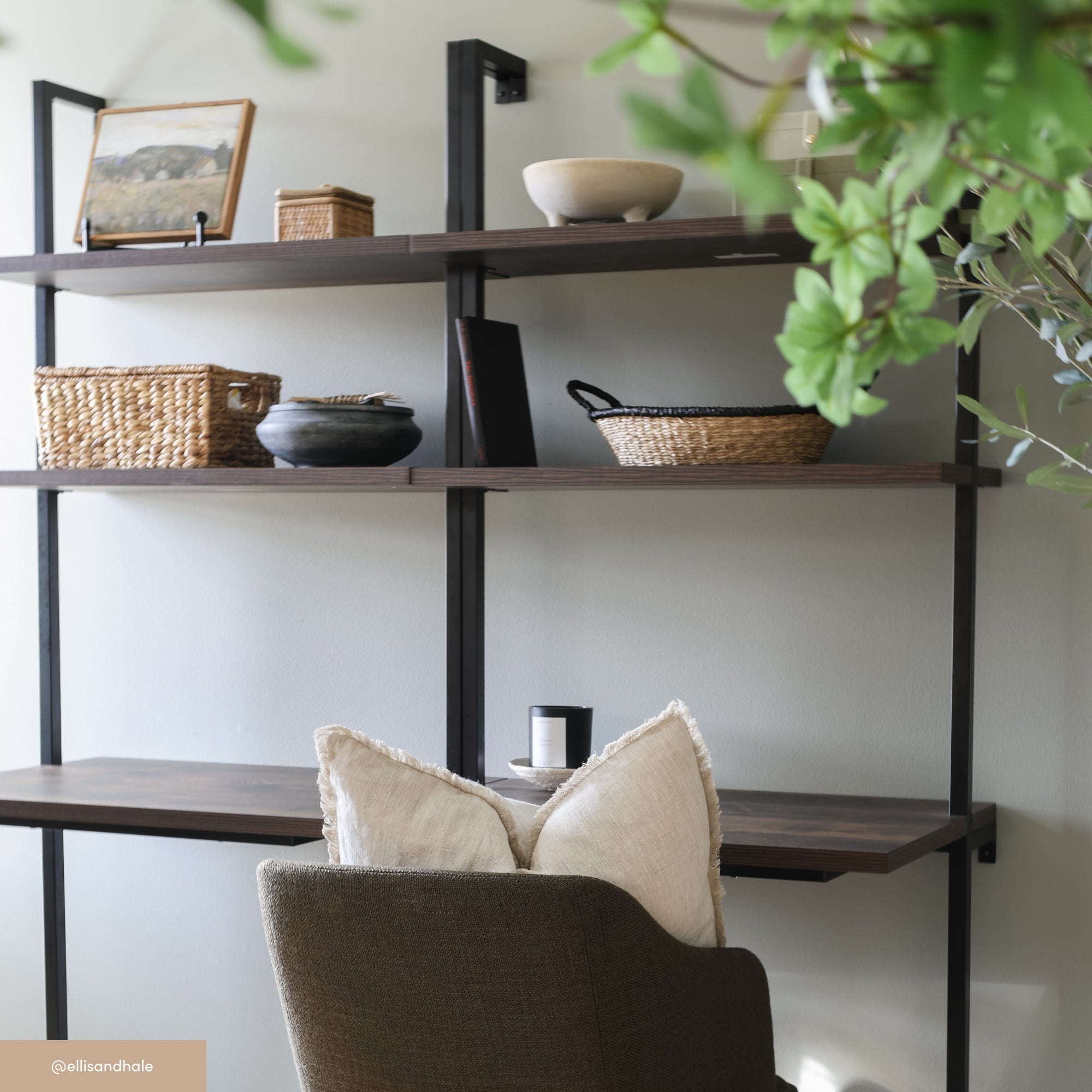 A home office corner with a brown chair, two cream pillows, and a wooden desk. Above the desk, open shelves hold baskets, bowls, a framed picture, and decorative items. Green leaves partially frame the scene.