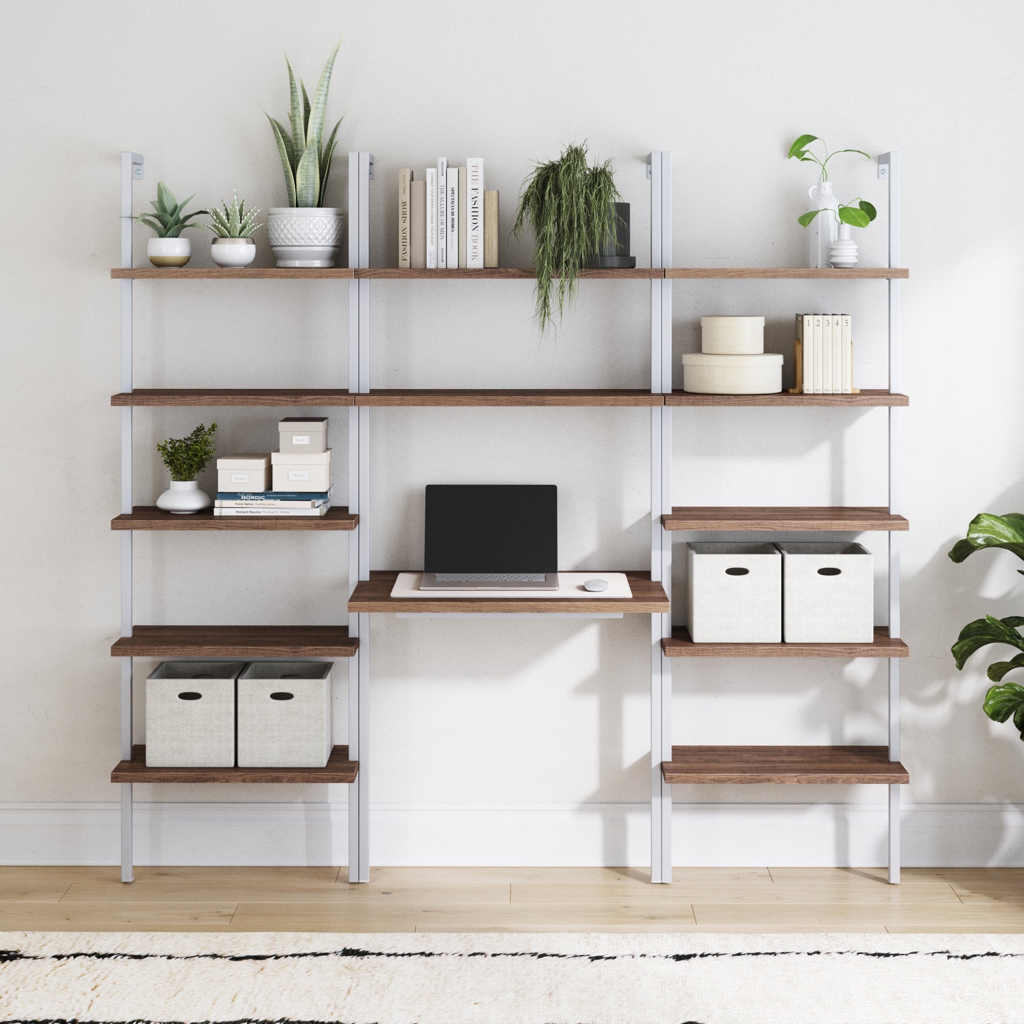 The Nathan James Wood & Metal Wall Mount Office Desk Set in Rustic Oak features ladder shelves that hold books, potted plants, storage boxes, and a closed laptop, creating a minimal, organized workspace with greenery against a white wall.