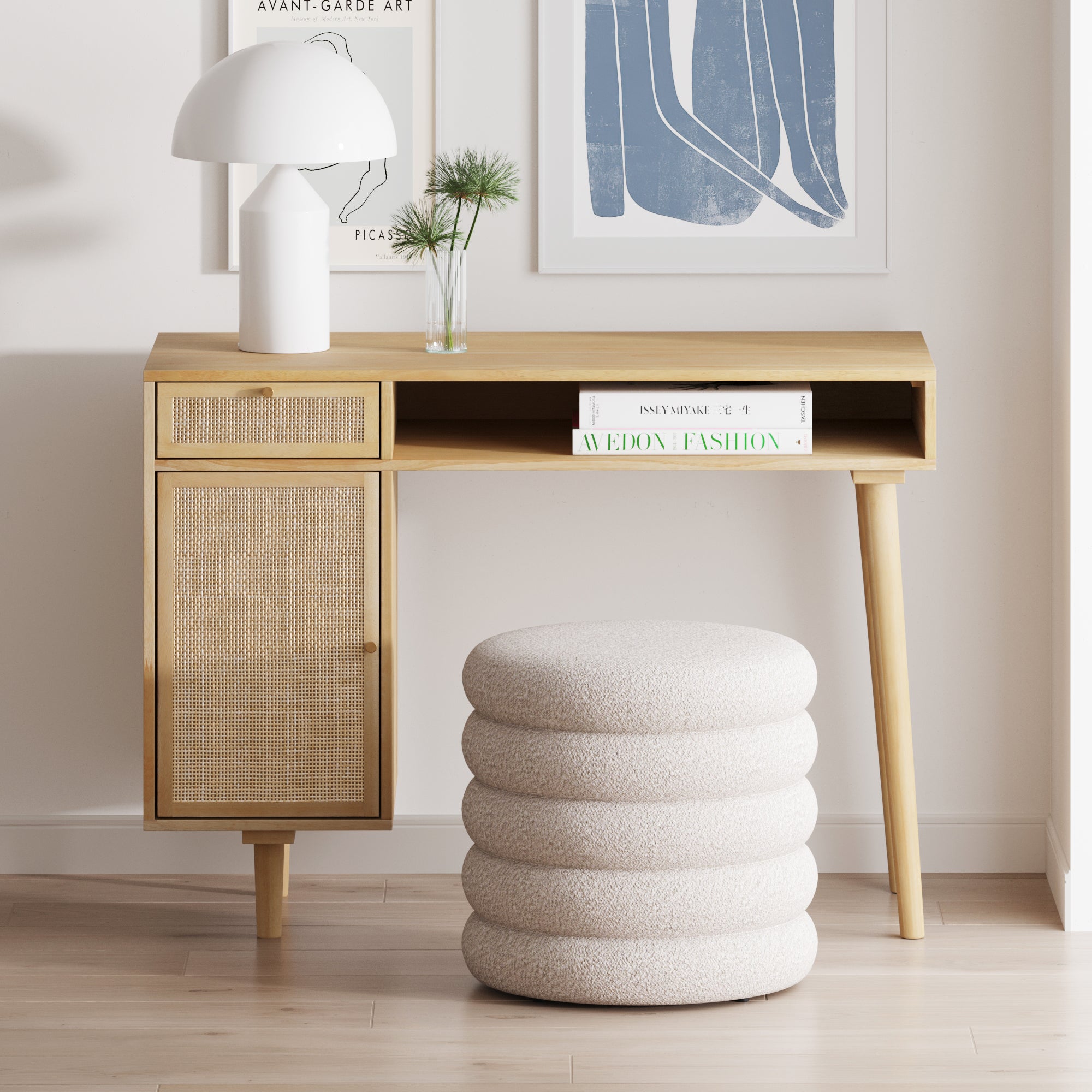 A minimalist wooden desk with a textured cabinet, a modern white lamp, two books, a small vase with plants, and a round, cushioned stool in a bright room with abstract art on the wall.