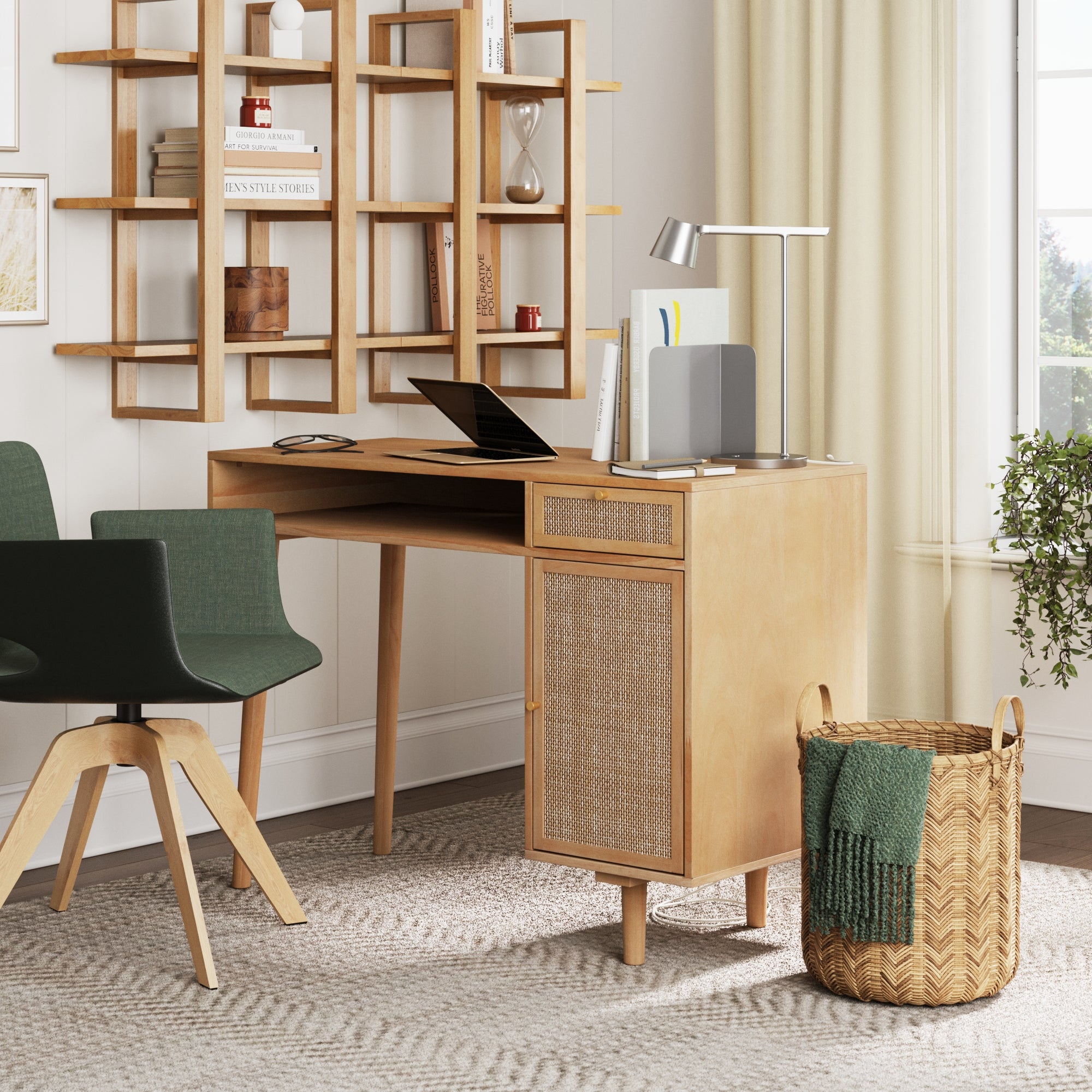 A modern home office features the Nathan James Solid Wood & Rattan Desk and Bookshelf Wall Unit, a green chair, laptop, lamp, and notebooks. Books and decor fill the shelves. A woven basket with a green blanket sits on a beige rug by cream curtains.