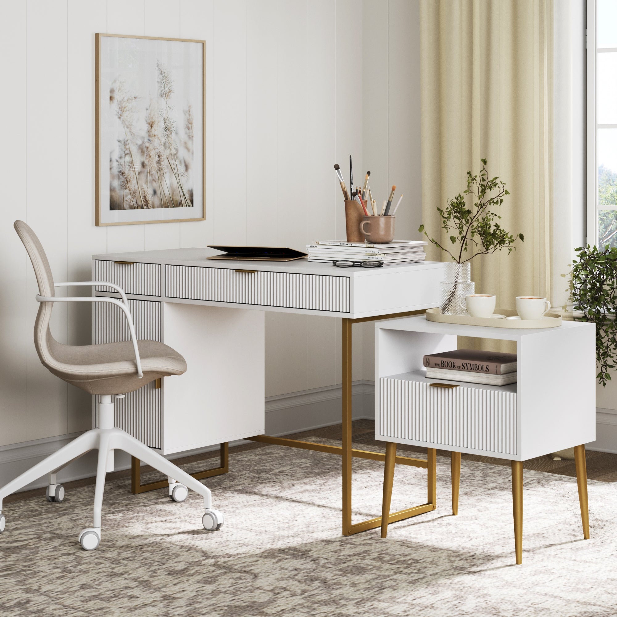 A modern home office features the Nathan James Fluted Desk & Nightstand Bedroom Vanity Set, a beige office chair, books and a plant on the nightstand, art supplies on the desk, framed wall art, and beige curtains by the window.