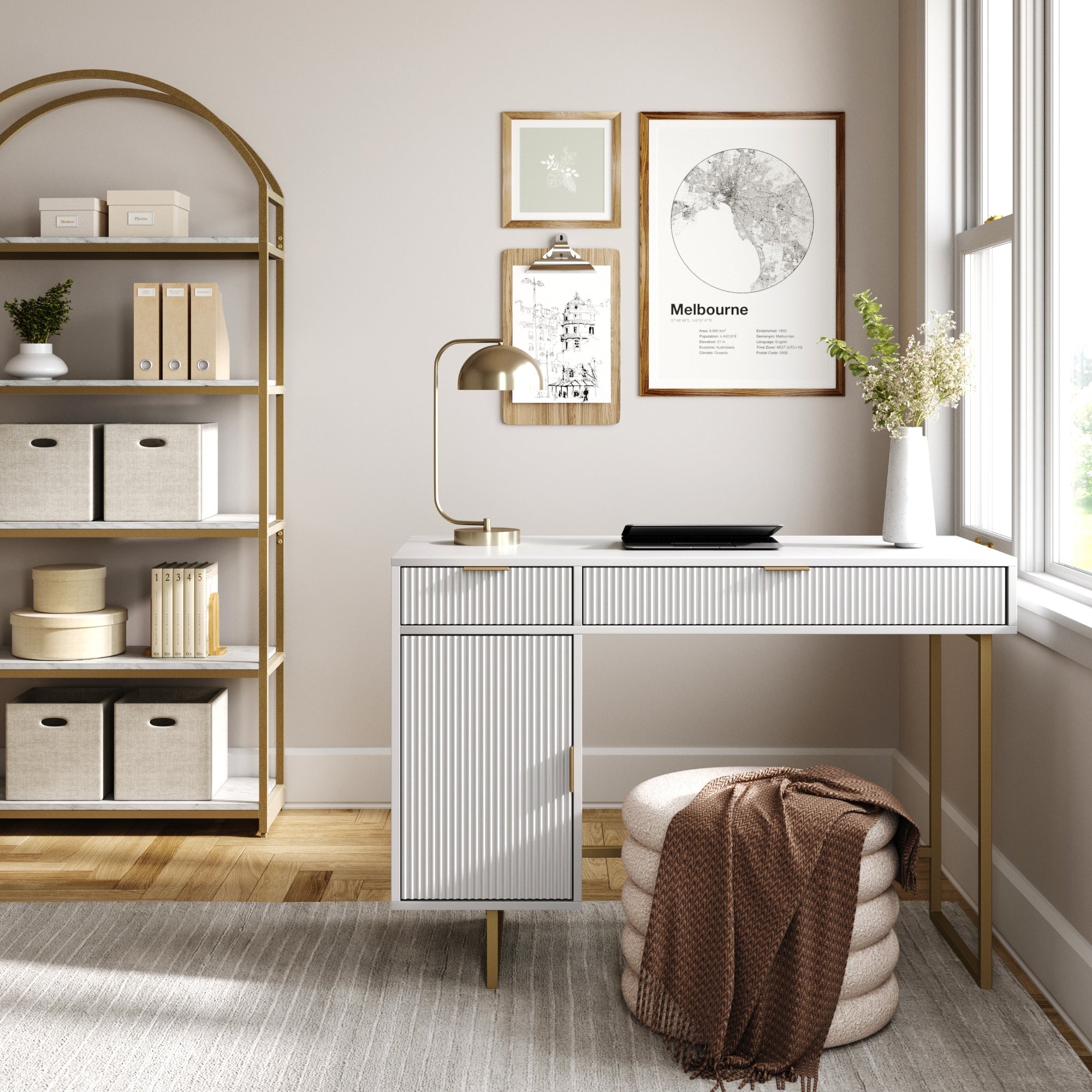 A modern minimalist home office with the Nathan James Mid-Century Fluted Storage Vanity Desk, gold lamp, framed wall art, bright window, shelving unit for storage boxes, and a beige pouf draped in a brown blanket.
