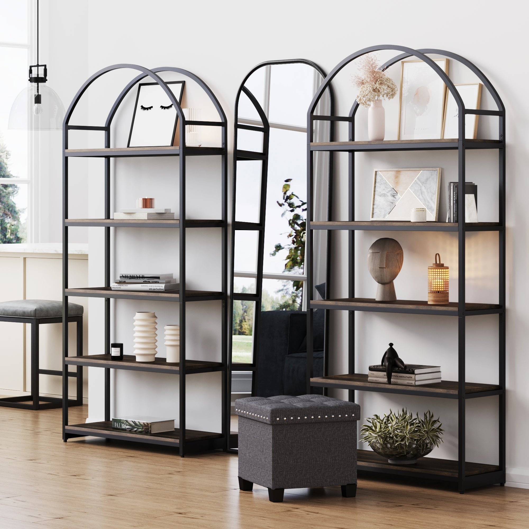 Two Nathan James Metal 5-Shelf Arched Bookshelves in black stand beside a tall mirror, styled with books, plants, lamps, and decor on their nutmeg-finish shelves. A gray ottoman rests in front on a light wood floor.