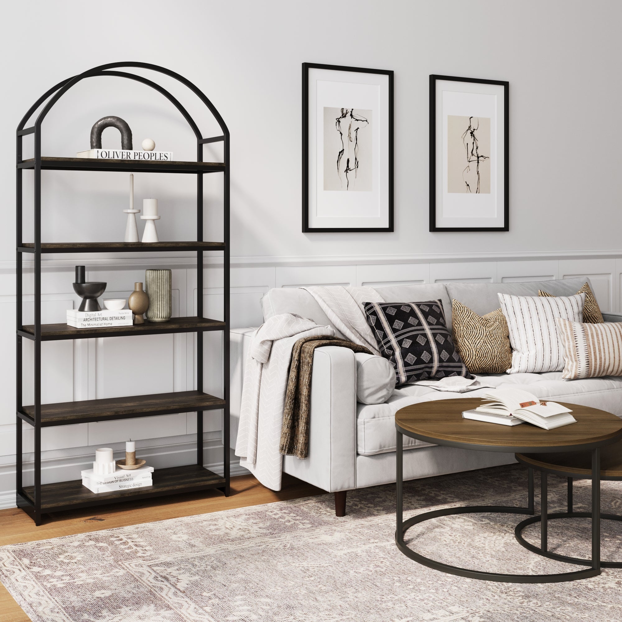 A modern living room with a light gray sofa, patterned pillows, a round coffee table, a black metal bookshelf with decor items, and two abstract art pieces on the wall. The space has a neutral, minimalist style.