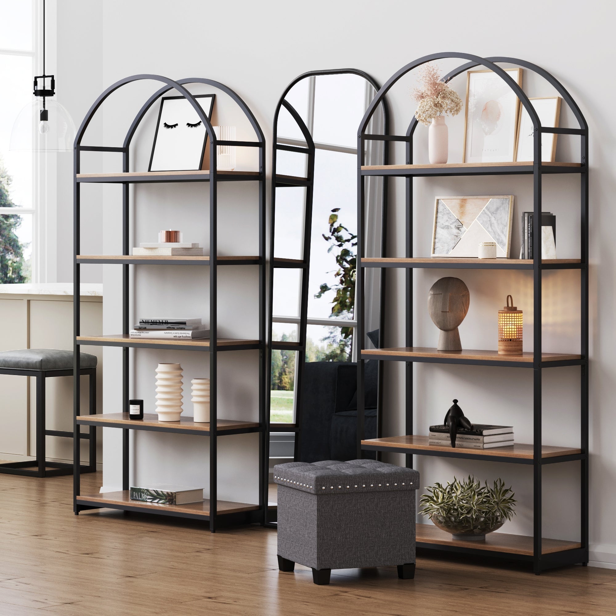 A pair of Nathan James Metal 5-Shelf Arched Bookshelves in oak, featuring matte black frames, stand against a white wall and display vases, books, art, and a lantern. Nearby are a window, mirror, and gray ottoman.