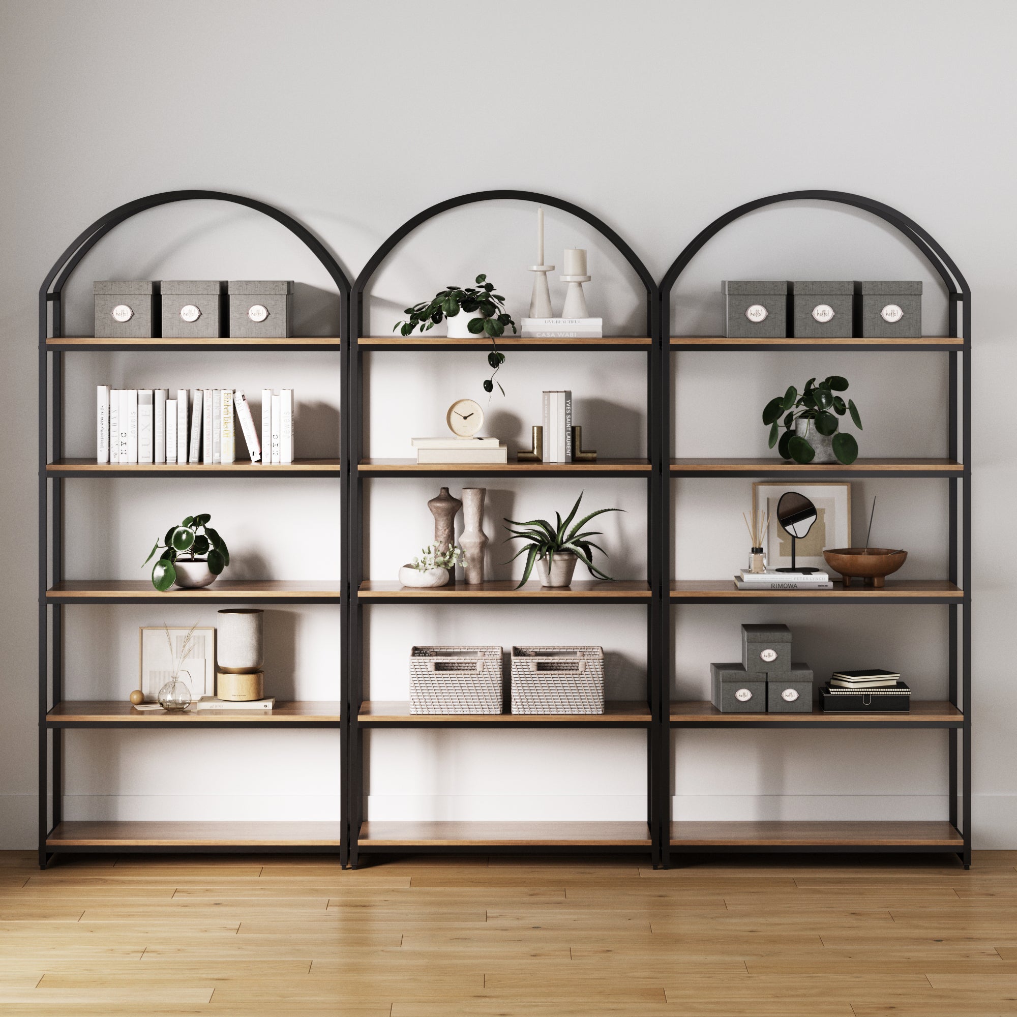 Three Nathan James Metal 5-Shelf Arched Bookshelves in oak (set of 3) stand against a white wall, neatly displaying books, gray storage boxes, plants, decor, and baskets on a light wood floor.
