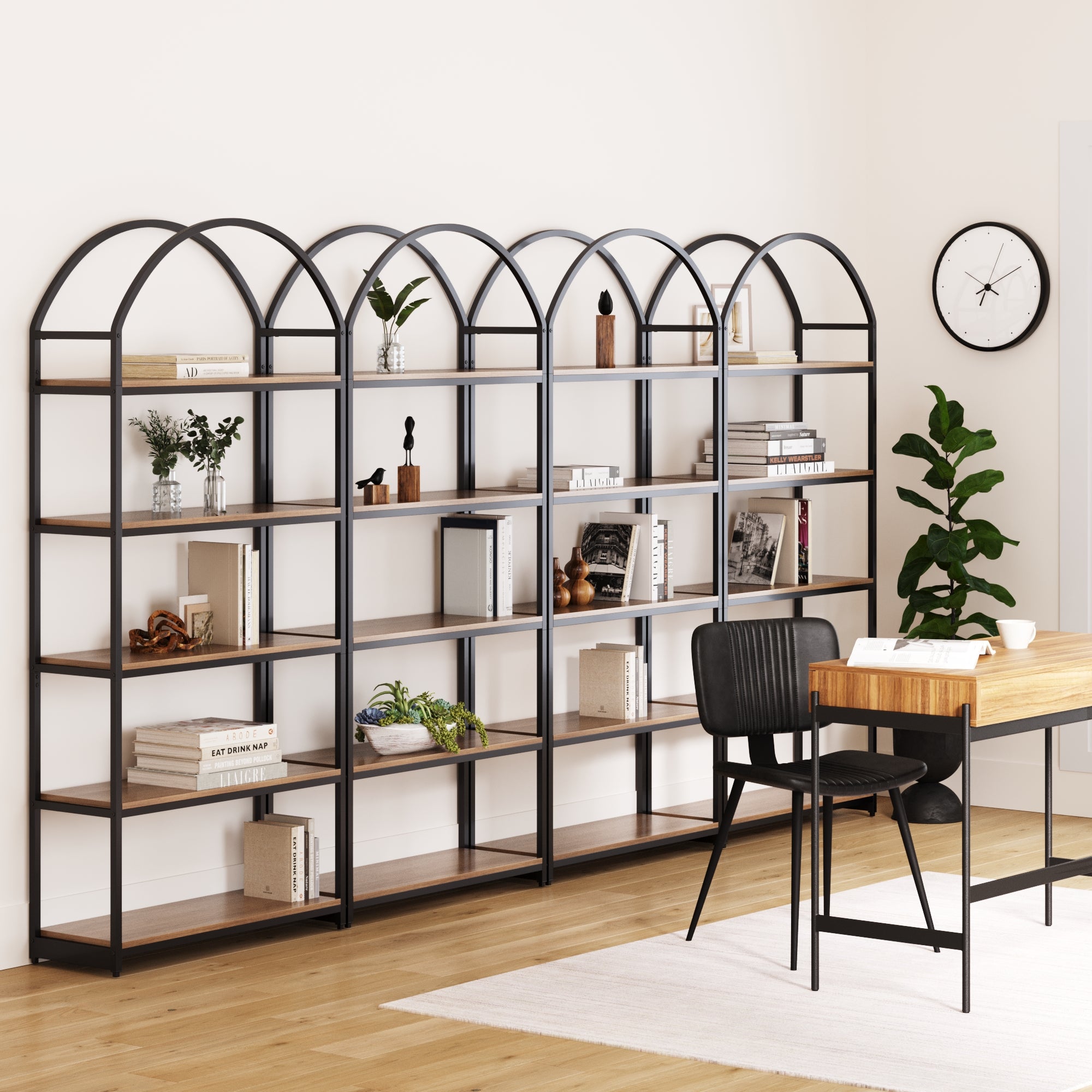 A modern home office features four Nathan James Metal 5-Shelf Arched Bookshelves in oak, displaying books, plants, and decor. A wooden desk with a black chair sits nearby amid light walls, a plant, wall clock, and wooden floors.