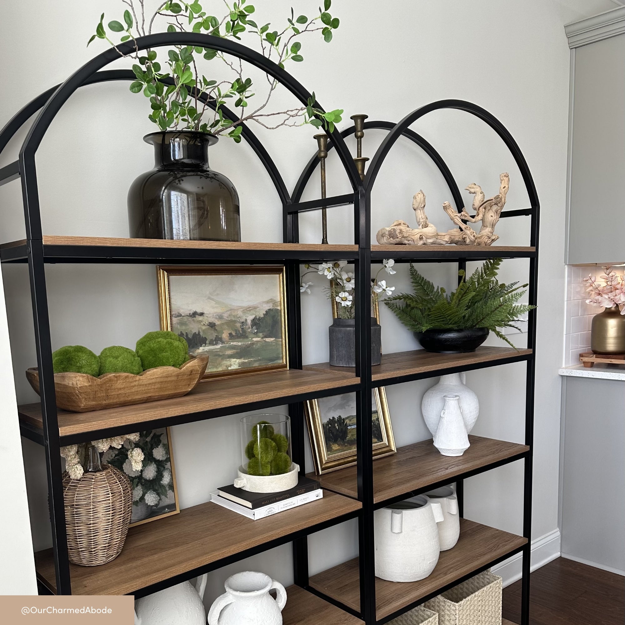 A black metal and wood bookshelf displays decorative items: vases, framed art, green plants, faux moss balls, baskets, and ceramic pitchers. The shelf is styled in a modern, minimalist living space with neutral tones.