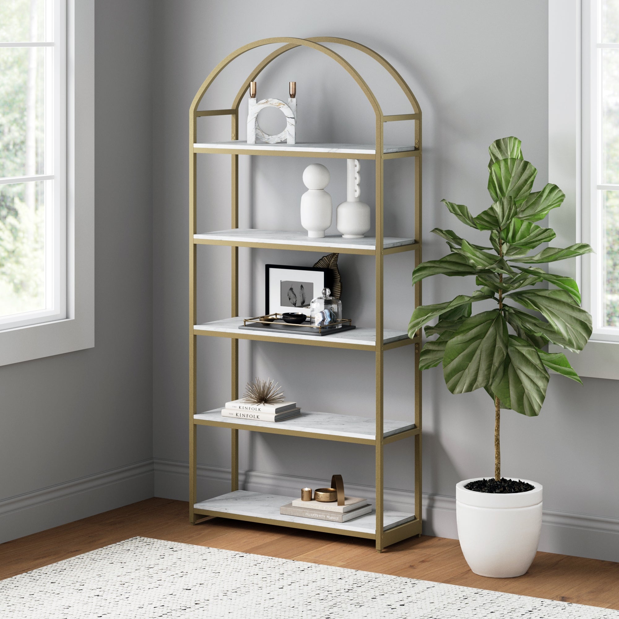 A gold and white arched bookshelf in a corner holds modern decor items. A potted plant in a white pot sits on the wood floor next to it. Two windows let in sunlight, brightening the gray-walled room.