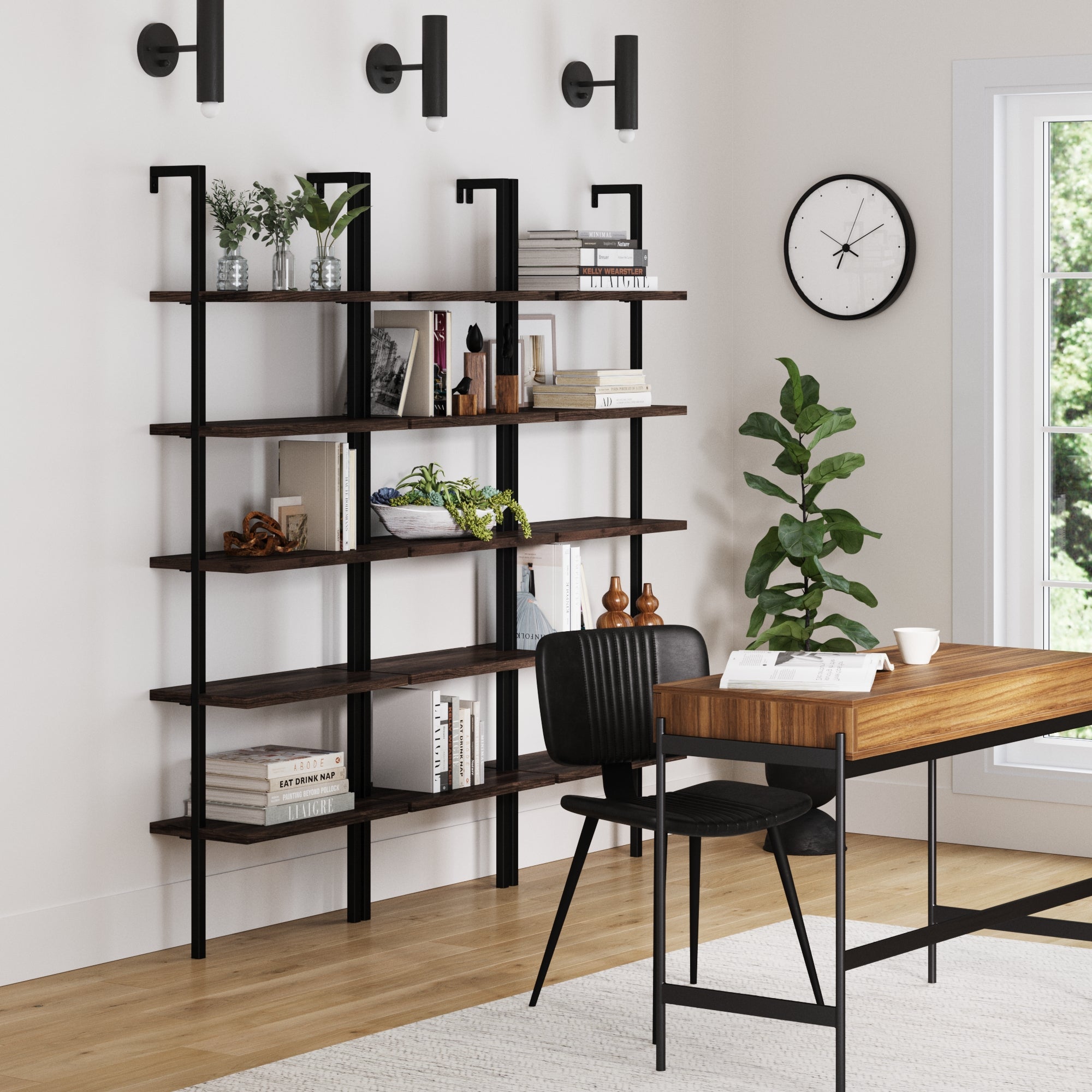 Modern office with a wooden desk, black chair, and wall clock. Three Nathan James 5-Shelf Ladder Wall Bookshelves in Nutmeg display books, plants, and decor beneath black wall lights. A large window and potted plant are on the right.