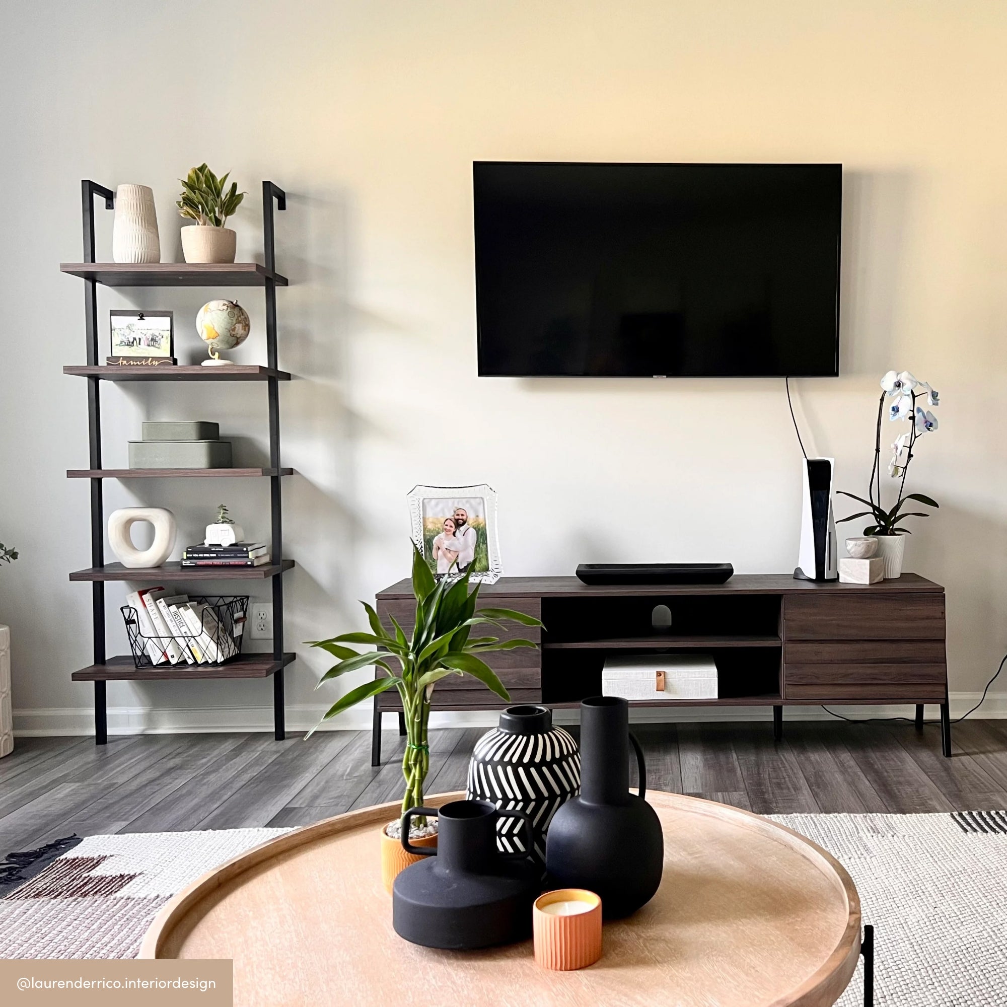A modern living room with a wall-mounted TV, a dark wood TV stand, a black metal and wood bookshelf, decorative plants, vases, framed photos, and a round coffee table with black and white ceramics in the foreground.