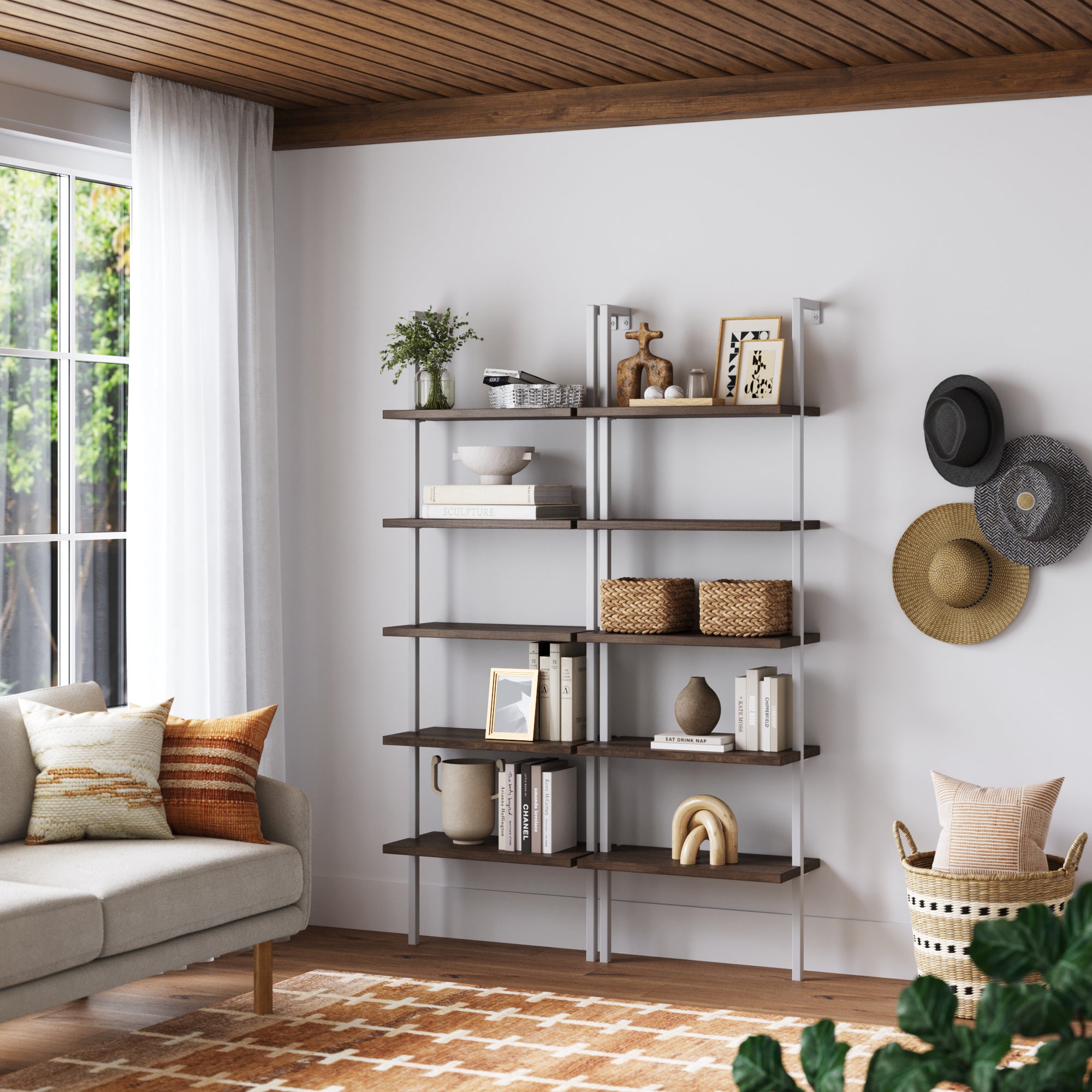 A modern living room features two Nathan James Ladder Bookshelf Oak Matte White sets holding books, baskets, plants, and decor. Nearby are hats on the wall, a cushioned sofa, woven basket, large window, and a patterned rug.