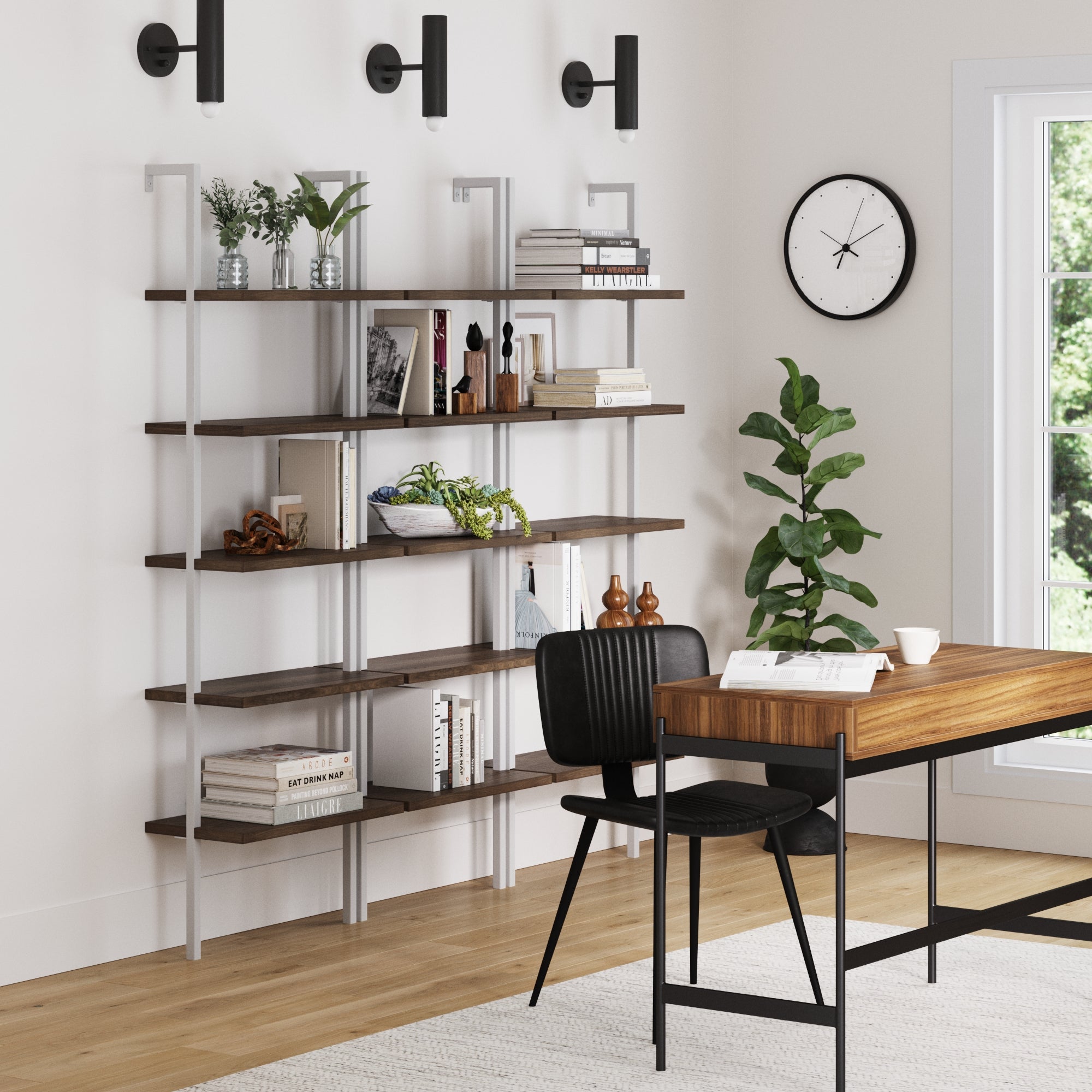 Modern home office with a wooden desk, black chair, Nathan James Ladder Bookshelf Oak Matte White Set of 3 filled with books and decor, wall clock, potted plant, three black wall lights, and a window letting in natural light.