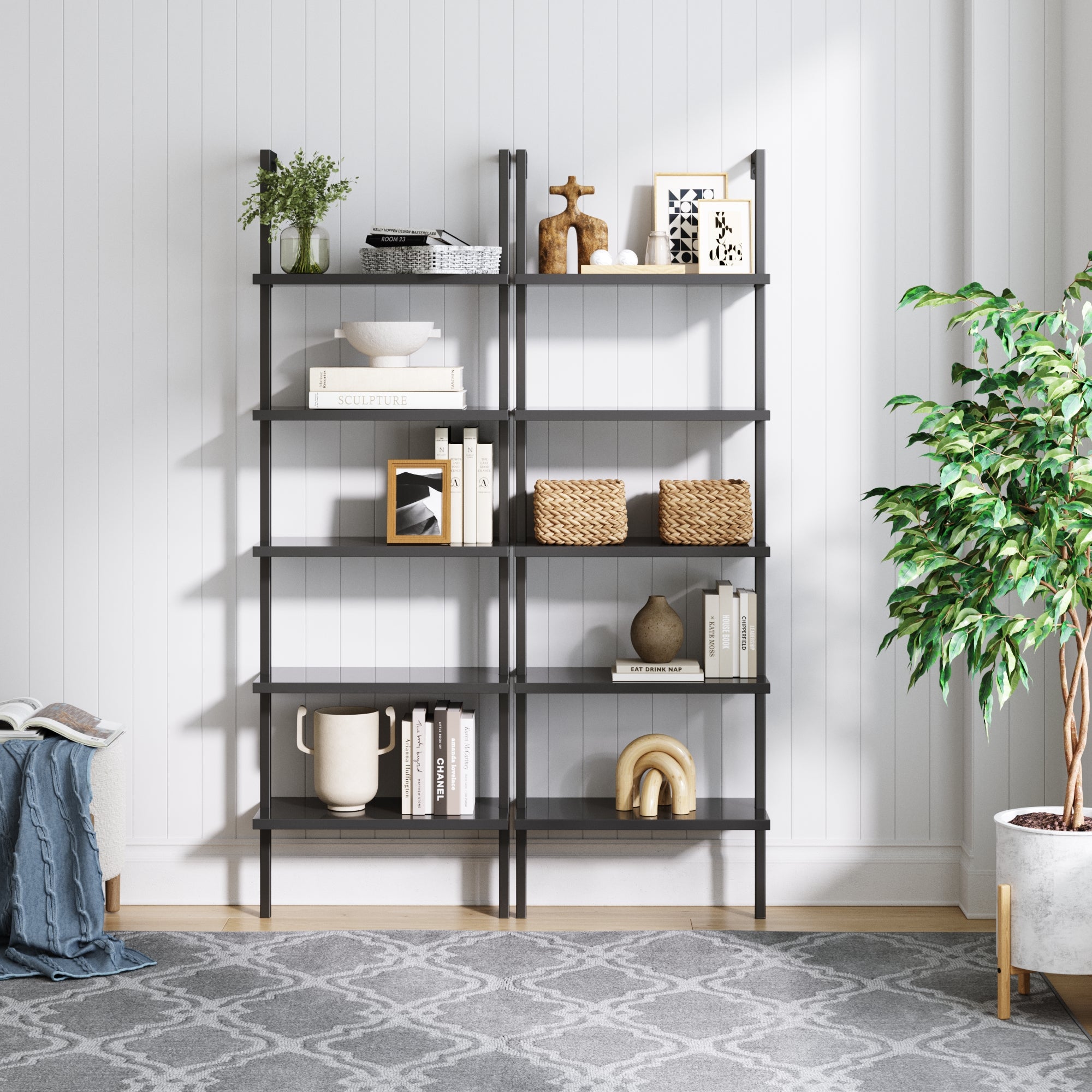 A modern black metal bookshelf holds books, decorative objects, baskets, and vases. It stands against a white paneled wall near a leafy green plant and a cozy chair with a blanket on a patterned gray rug.