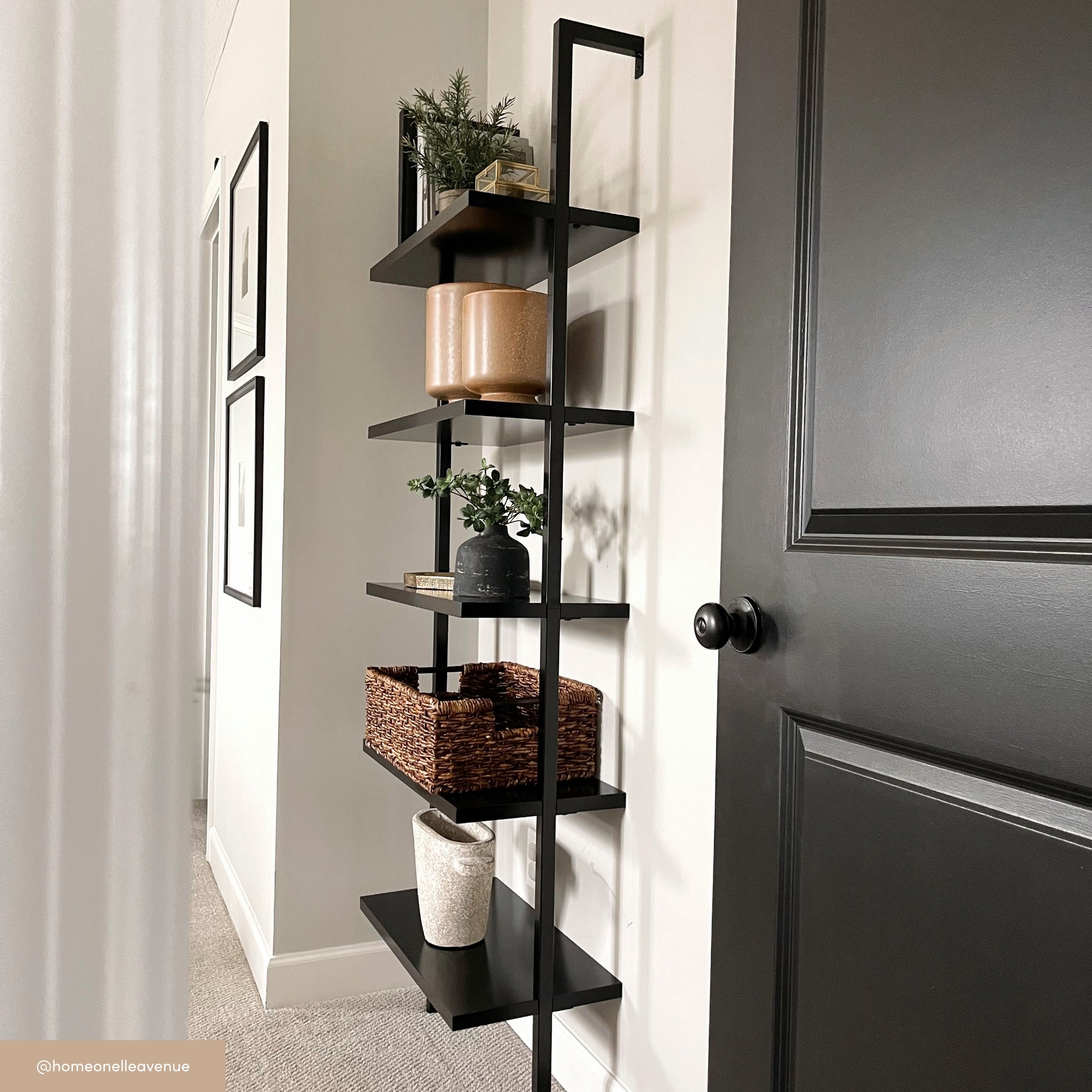 A black metal wall-mounted shelf holds decorative items including potted plants, a woven basket, vases, and a candle. The shelf is against a white wall near a black door in a modern, minimal room.