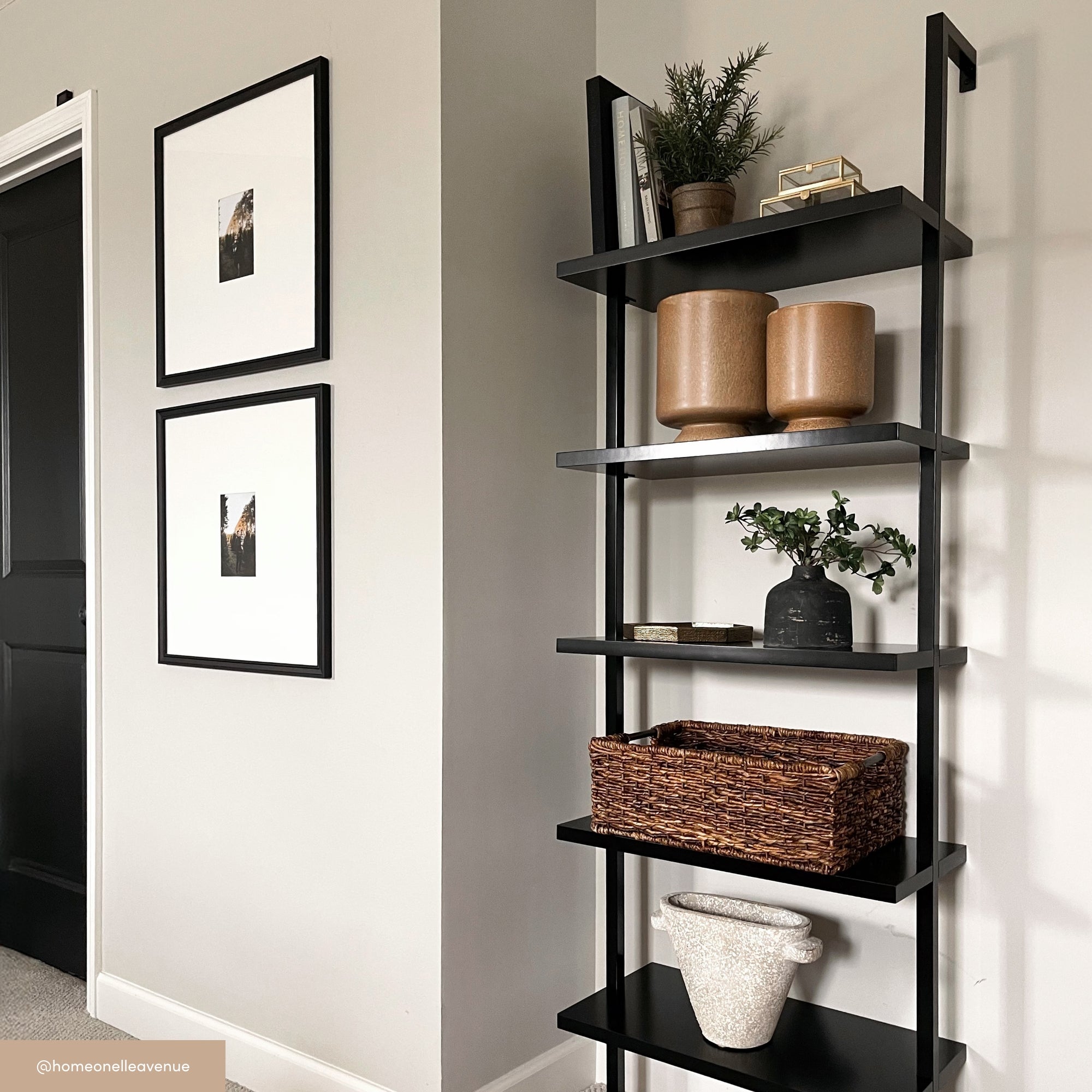 A black ladder-style shelf holds tan vases, a basket, a potted plant, and decor in a neutral-toned room. Two framed photos hang on the nearby wall, and a black door is partially visible.
