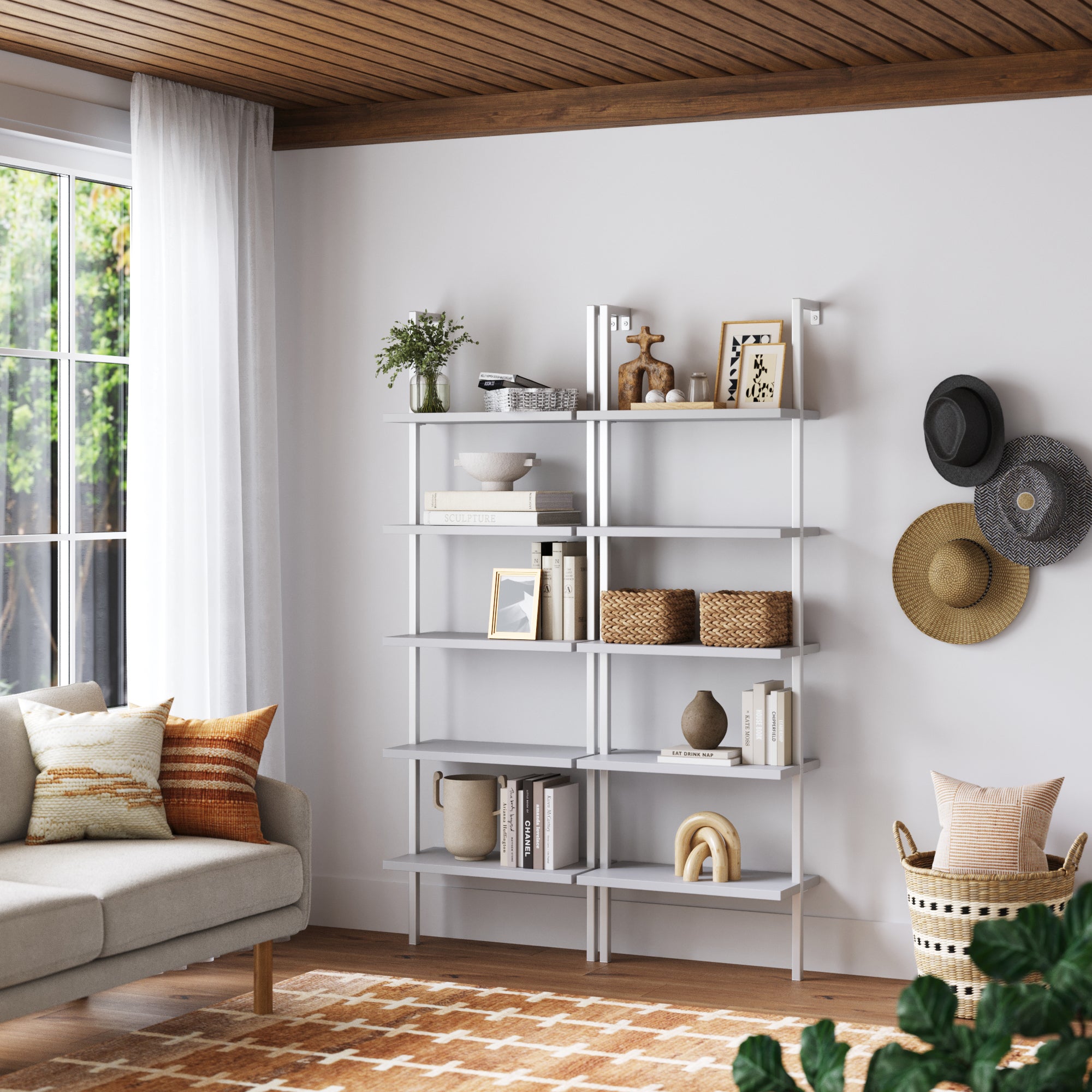 A modern living room features two Nathan James 5-Shelf Coastal Ladder Bookshelves in white displaying books, plants, baskets, and decor. A beige sofa with patterned pillows is by a large window with sheer curtains, and three hats hang on the wall.