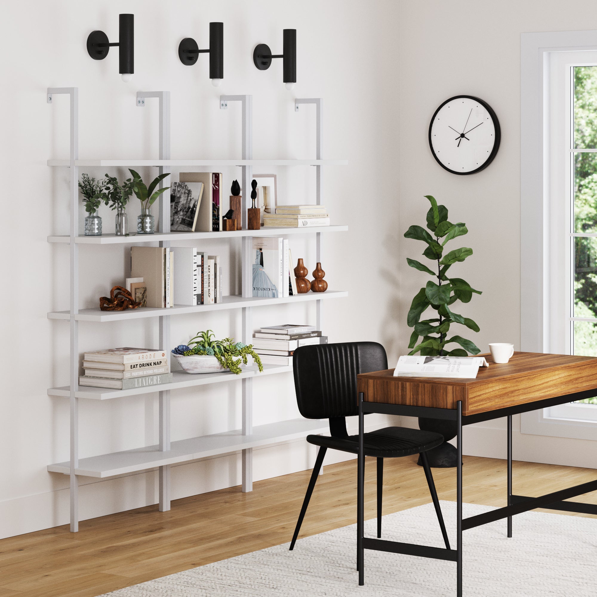 Modern home office with a wooden desk, black chair, and three white Nathan James 5-Shelf Coastal Ladder Bookshelves filled with books and decor, complemented by black wall sconces, a clock, potted plant, and sunlight streaming in.