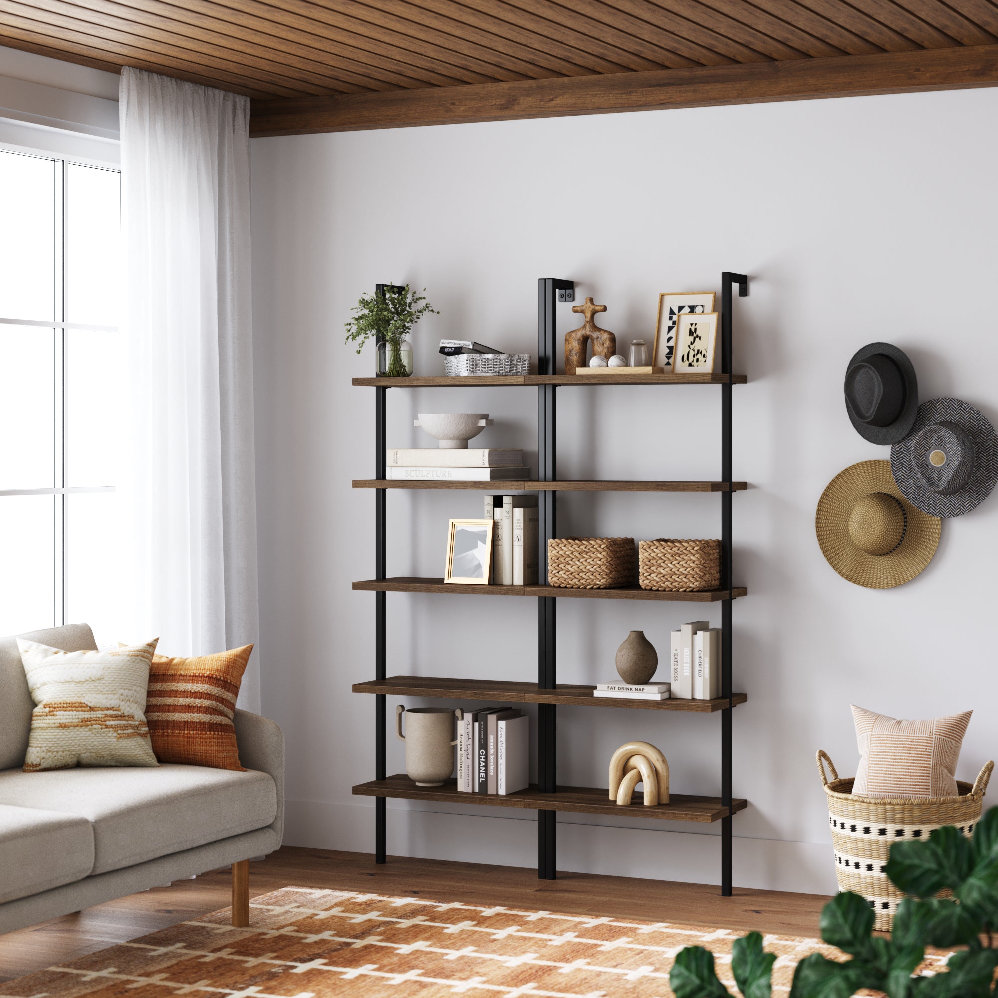 A modern living room features the Nathan James 5-Shelf Ladder Bookshelf Oak Matte Black (Set of 2), styled with books, baskets, and plants against a white wall. Straw hats hang nearby, with a beige sofa and patterned pillows on a geometric rug.