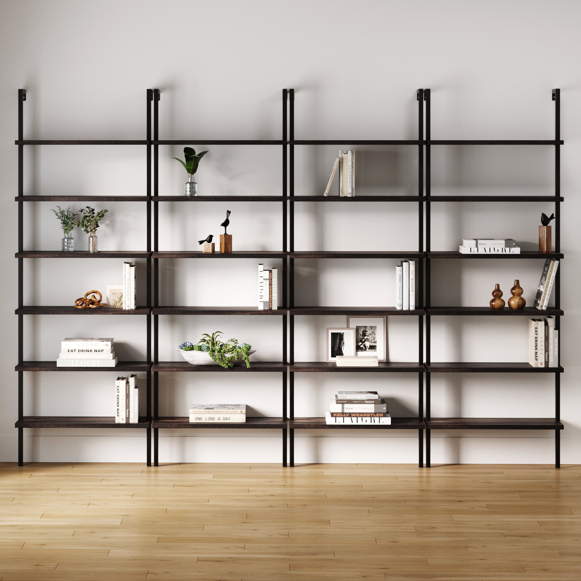 A set of four Nathan James 6-Shelf Bookcases in Nutmeg Matte Black stand against a plain white wall on light wood flooring, each sparsely decorated with books, small potted plants, vases, and decorative objects.