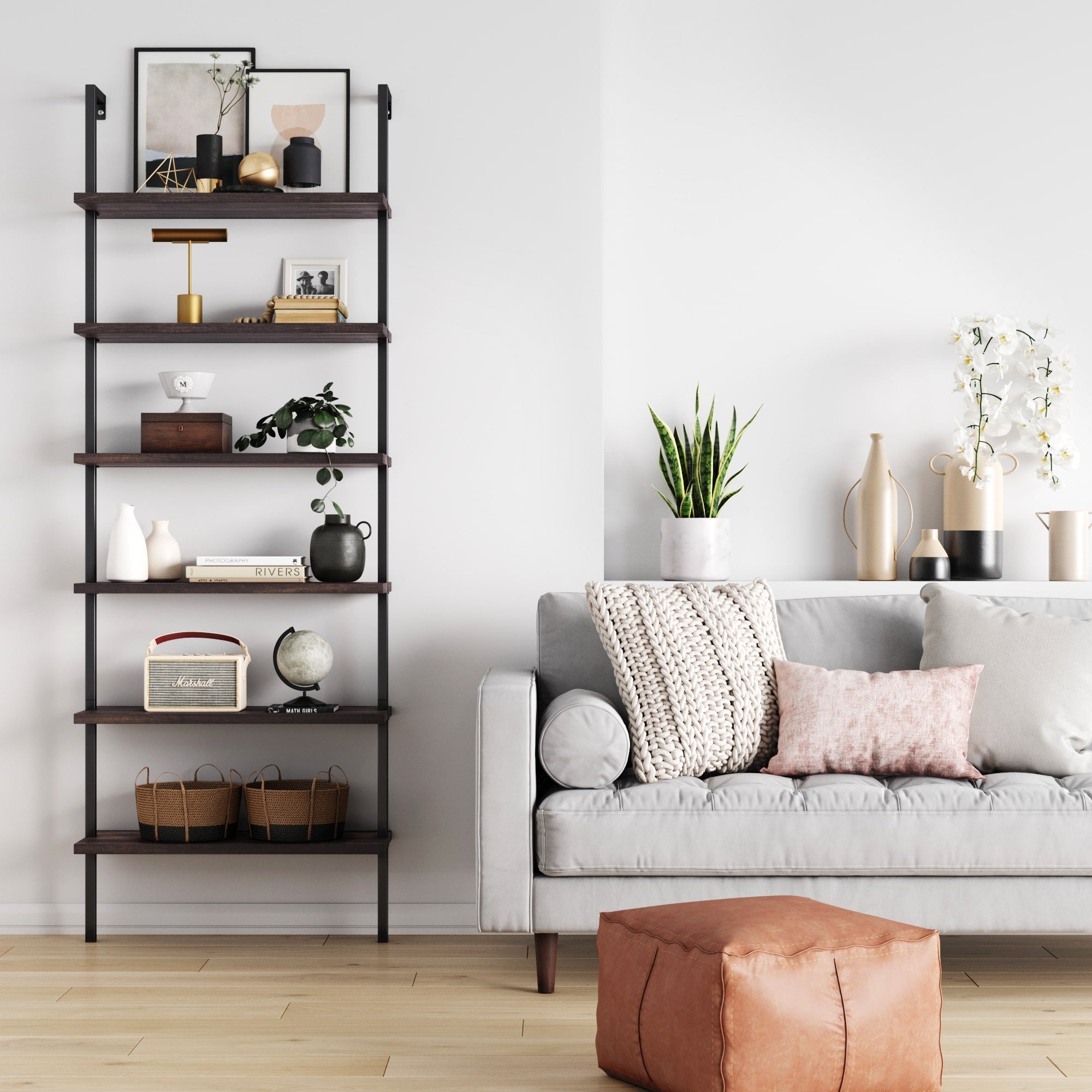 A modern living room features a gray sofa with pillows, a leather pouf, a tall dark wood bookshelf with decor items, potted plants, and a white console table holding vases and flowers against a white wall.