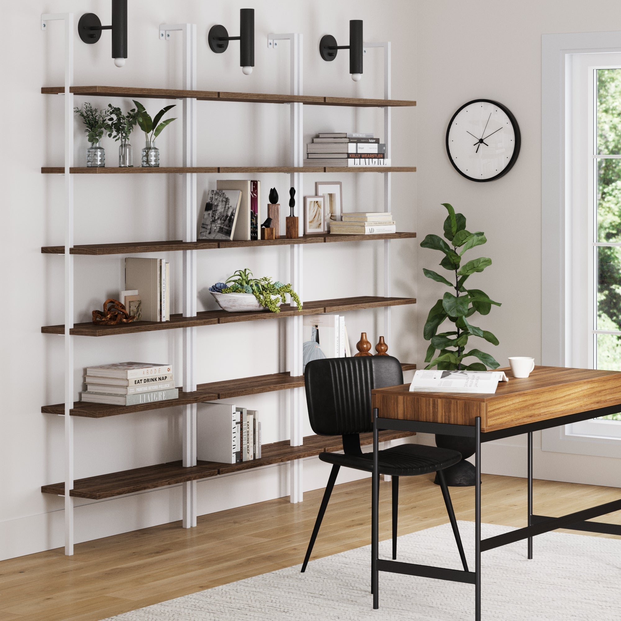 A modern home office features a wooden desk, black chair, and three Nathan James 6-Shelf Bookcase Oak Matte Whites displaying books and decor. A wall clock hangs above, while a potted plant by the window brightens the space.