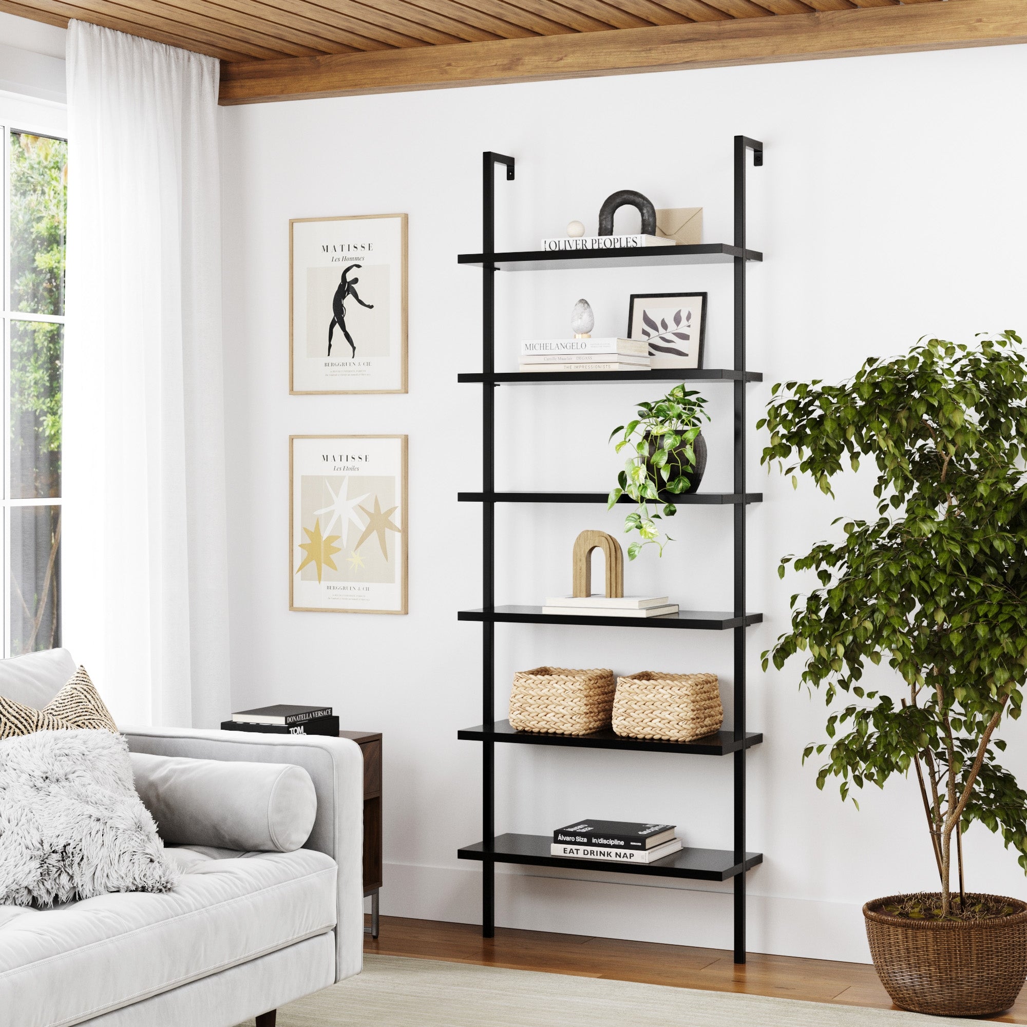 A modern living room features the Nathan James 6-Shelf Ladder Bookshelf in black displaying books, decor, and baskets. Set beside a large leafy plant, sunlight filters through white curtains into the neutral-toned space.