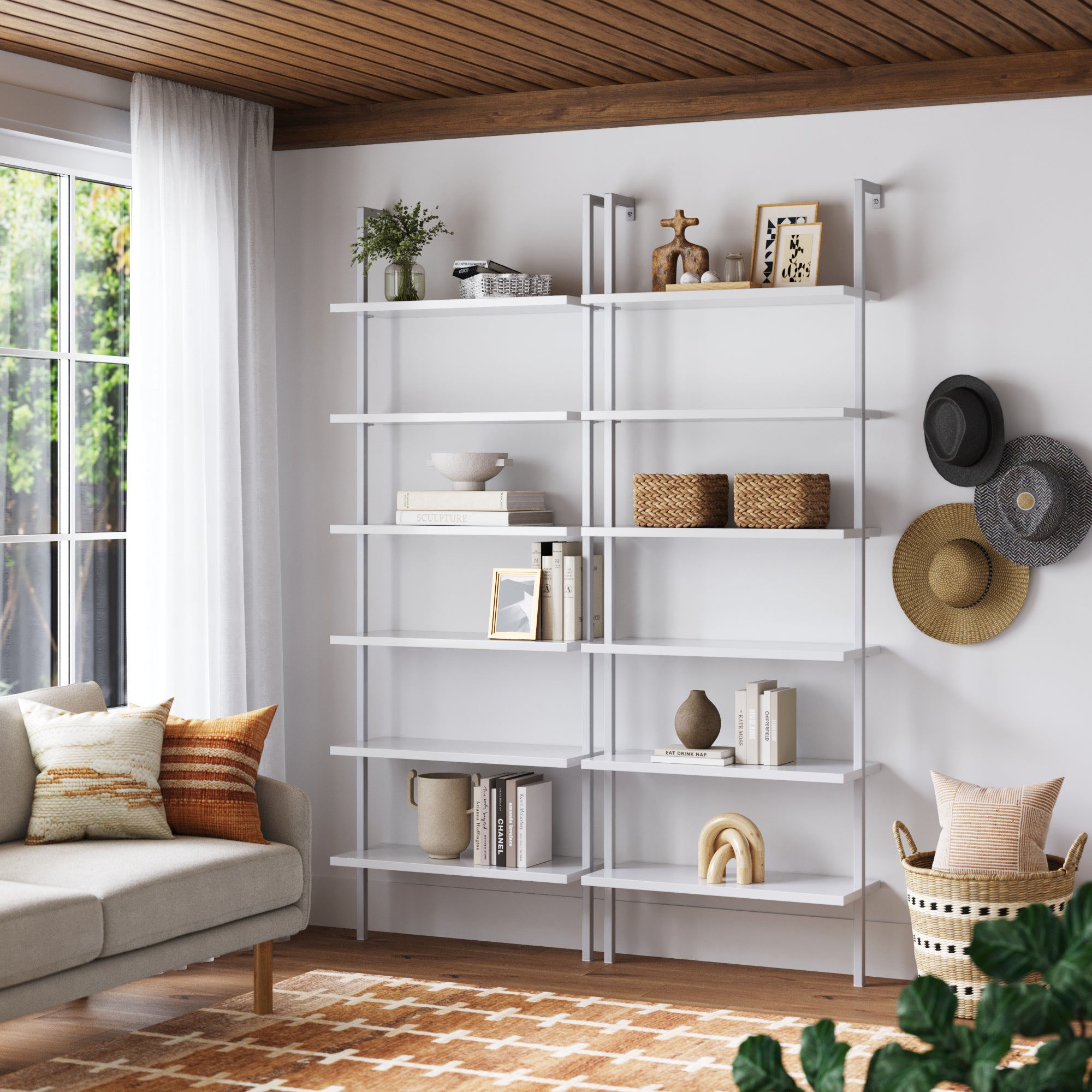 A modern living room showcases two Nathan James 6-Shelf Ladder Bookshelves in white, displaying books and decor. A beige sofa with patterned pillows, a patterned rug, and large windows with white curtains complete the space.