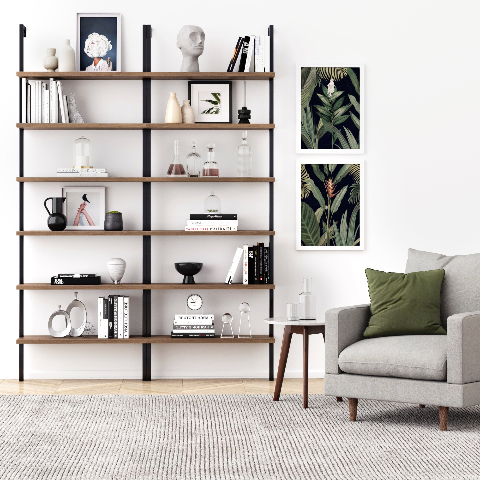 A modern living room with a gray armchair, green and white pillows, a small wooden side table, and a large bookshelf with decor, books, and glassware. Two botanical prints hang on the white wall.