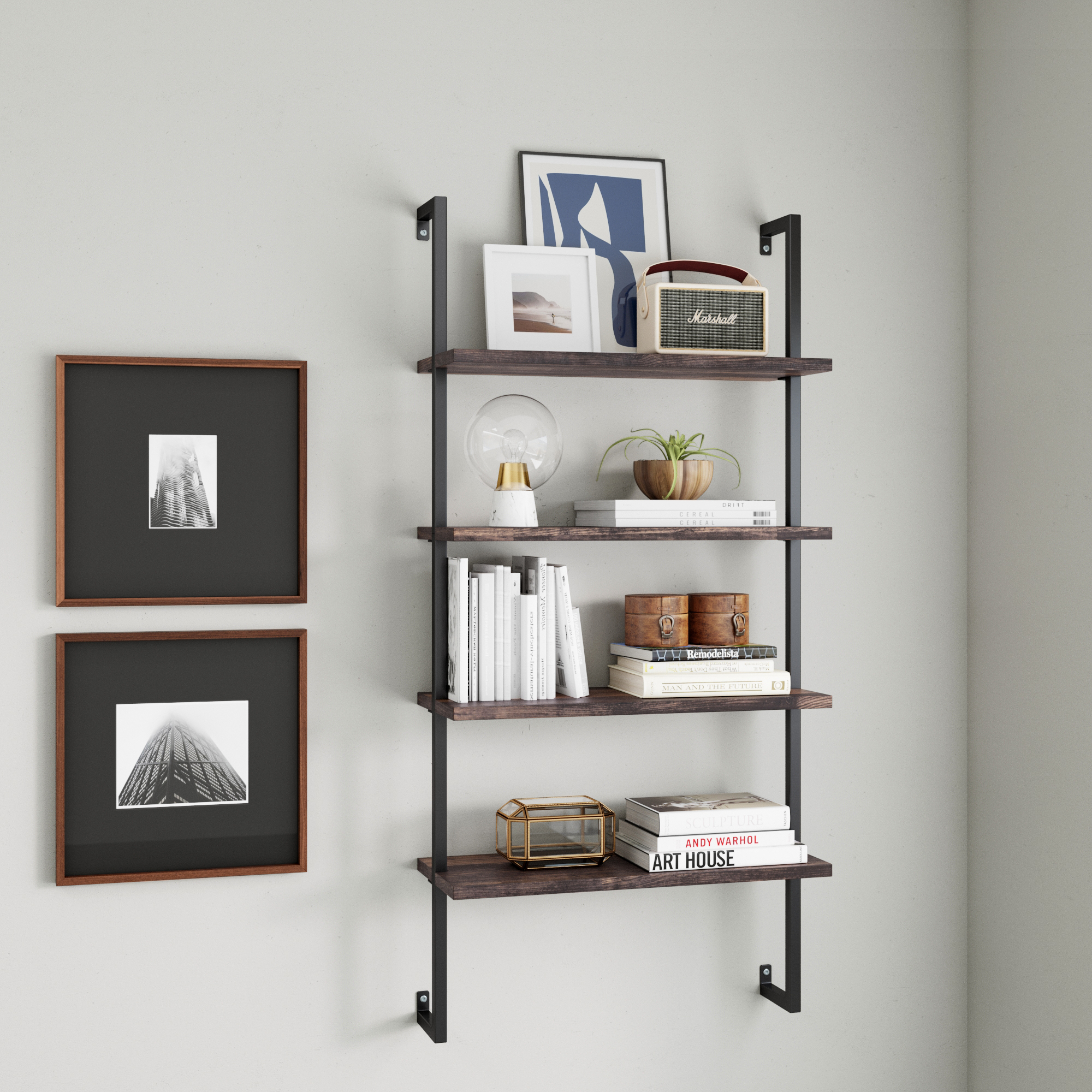 A wall-mounted bookshelf with three wooden shelves holding books, decorative items, a radio, a potted plant, and framed artwork, next to two black-and-white framed photos on a light-colored wall.