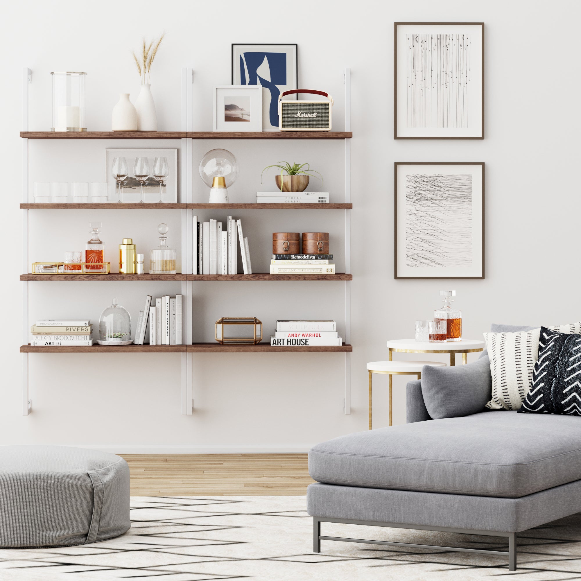 A modern living room with wall-mounted wooden shelves holding books, glassware, and decor, a gray sectional sofa, round ottoman, side tables, and framed art on white walls.
