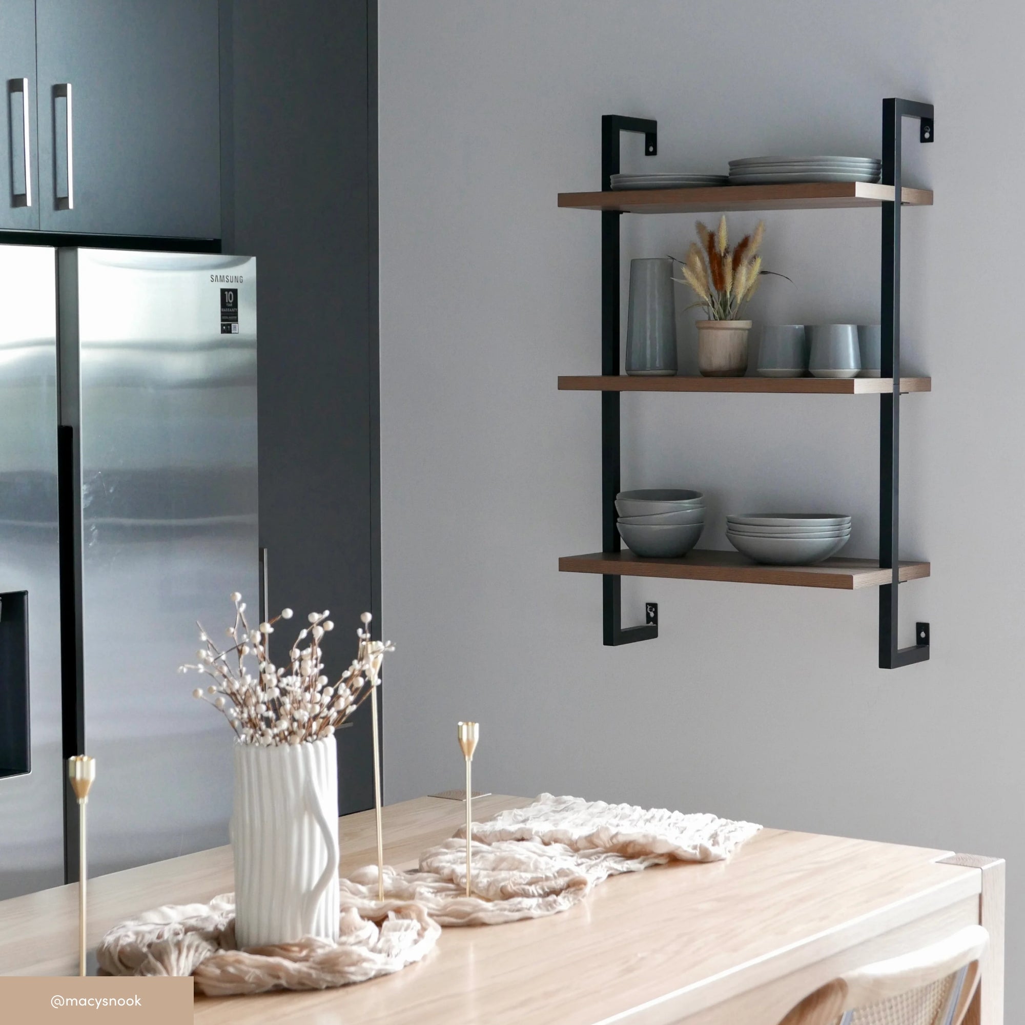 A modern kitchen with a light wooden table, decorative centerpiece, and three floating shelves on a gray wall holding plates, bowls, cups, and a vase with dried flowers; a stainless steel fridge is visible on the left.