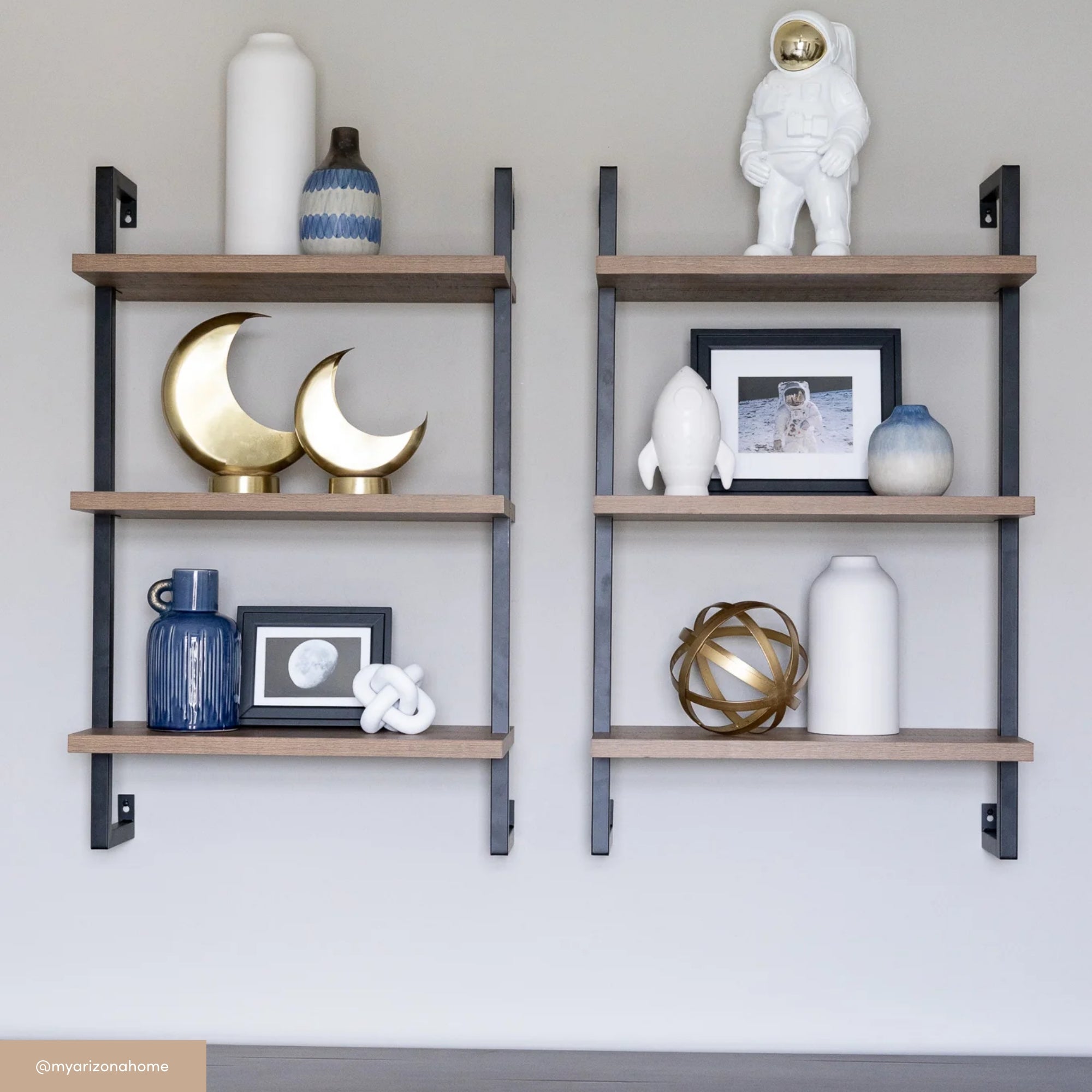 Two wall-mounted shelves display statues, vases, a blue bottle, gold crescent moons, a photo and figure of an astronaut, and decorative objects in white, blue, and gold tones against a light gray wall.